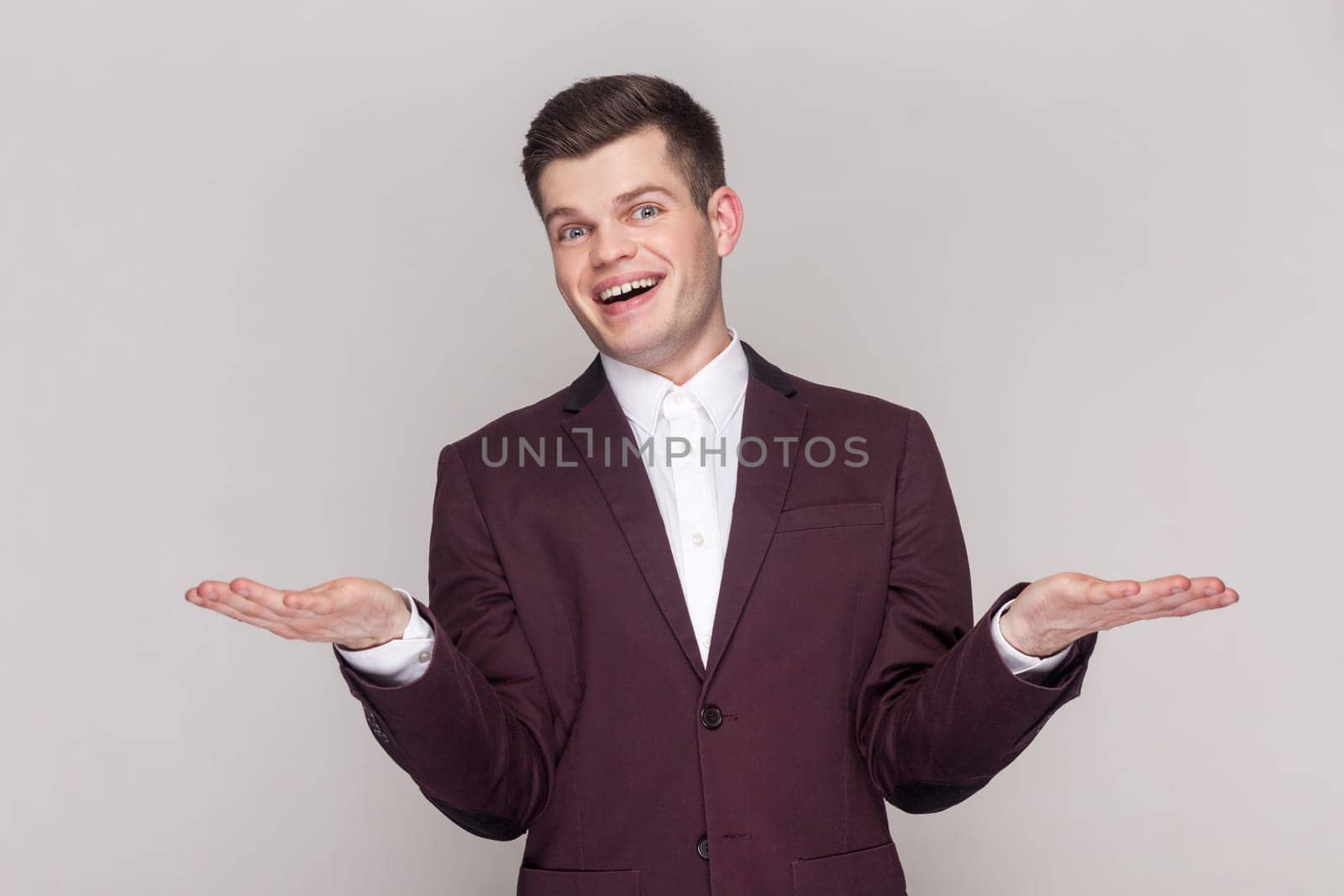 Portrait of pleased positive optimistic handsome man spreads hands, inviting guests, showing copy space on palms, wearing violet suit and white shirt. Indoor studio shot isolated on grey background.