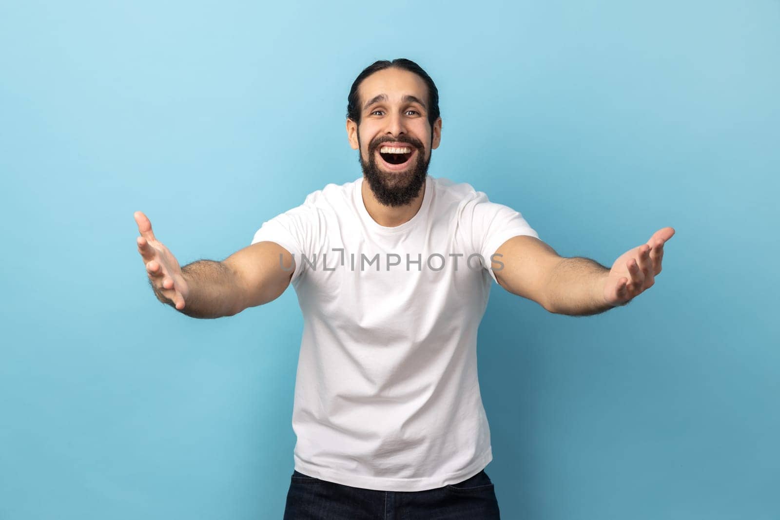 Welcome, wide open hug. Portrait of man with beard wearing white T-shirt stretching hands to camera and smiling broadly, going to embrace, share love. Indoor studio shot isolated on blue background.