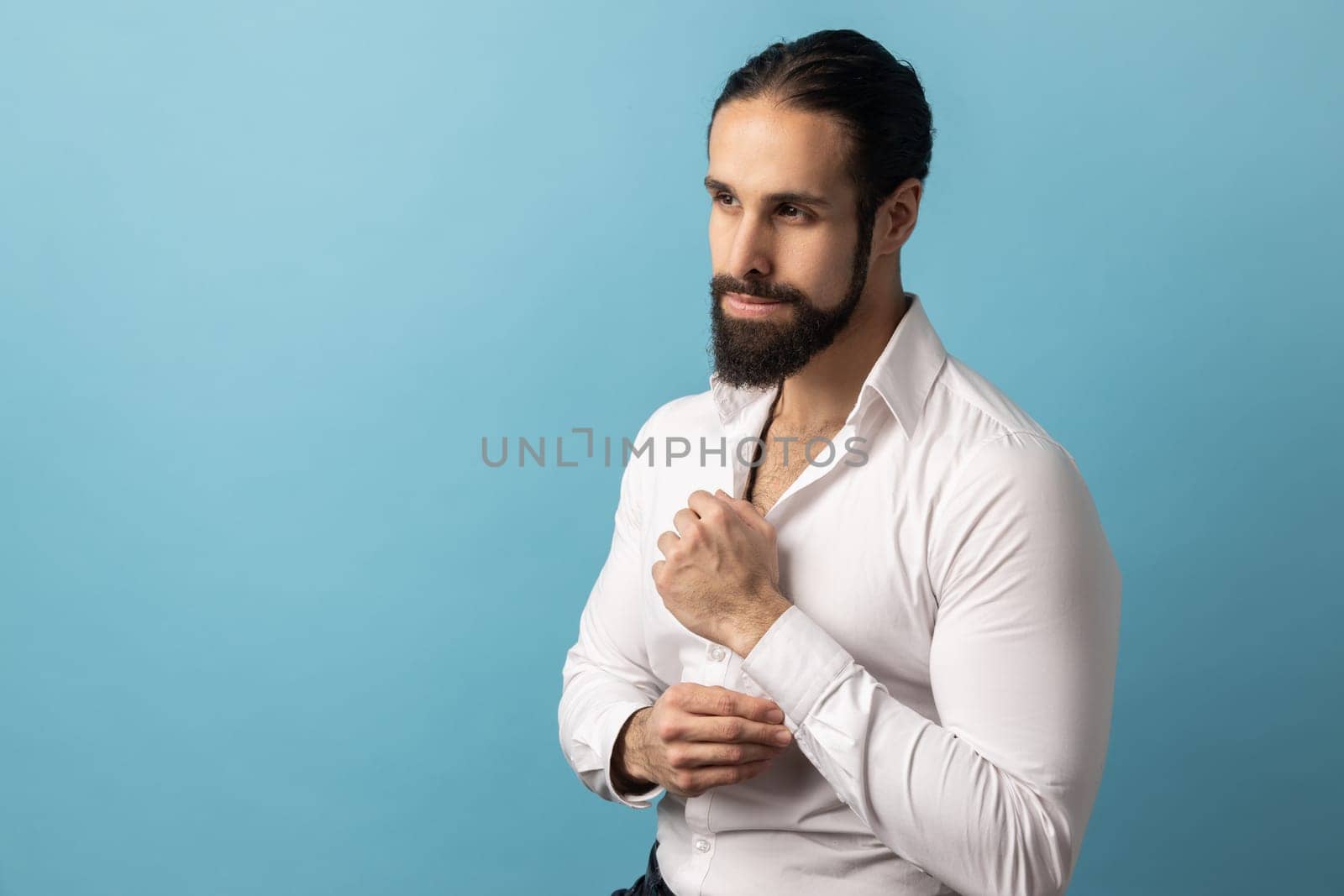 Portrait of handsome confident pensive man with beard wearing white shirt looking away and tucks cufflinks, going to an important event. Indoor studio shot isolated on blue background.