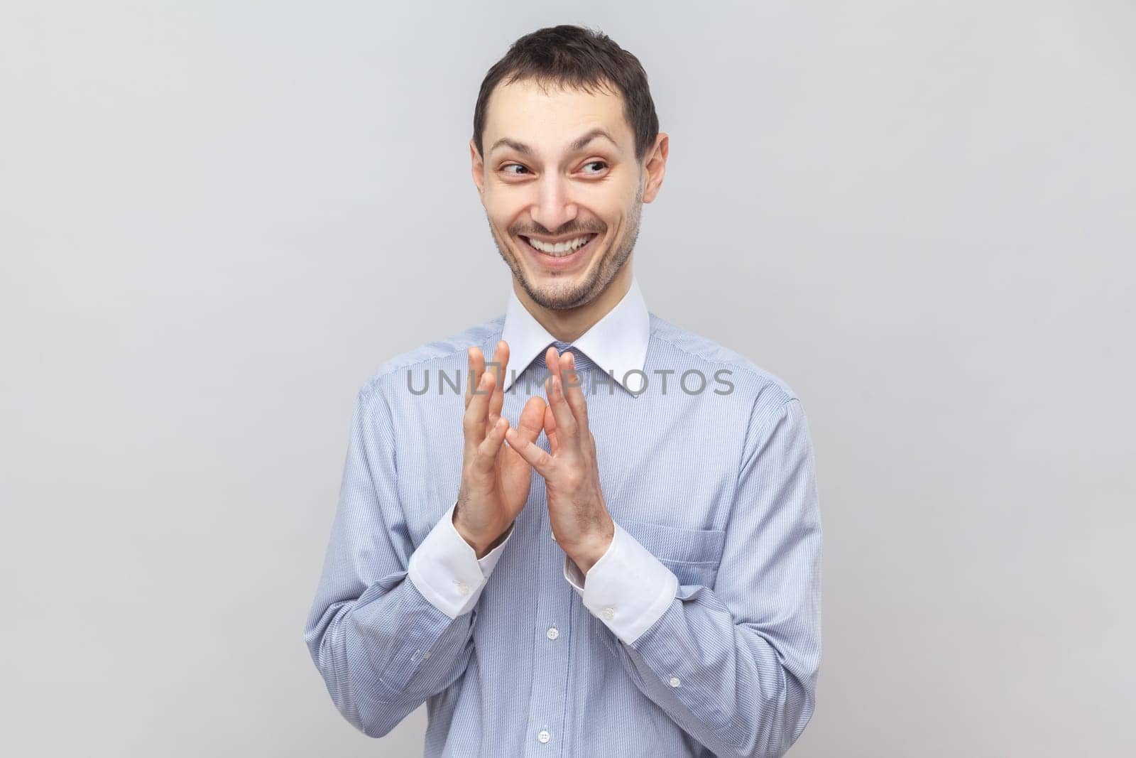 Portrait of cunning sly young adult man standing looking away with toothy smile, having devil plans, planning prank, wearing light blue shirt. Indoor studio shot isolated on gray background.