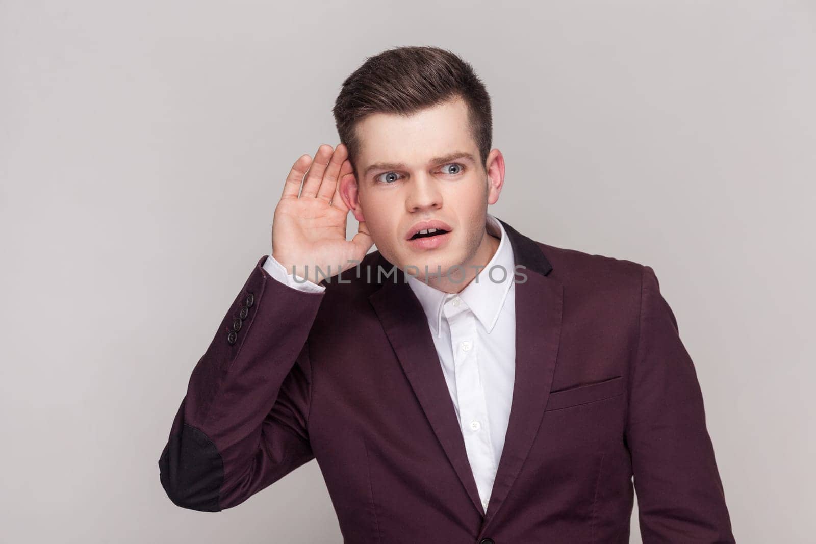 Young man holding hand near ear trying to hear secrets and find out gossips, listen to private whispers, , wearing violet suit and white shirt. Indoor studio shot isolated on grey background.