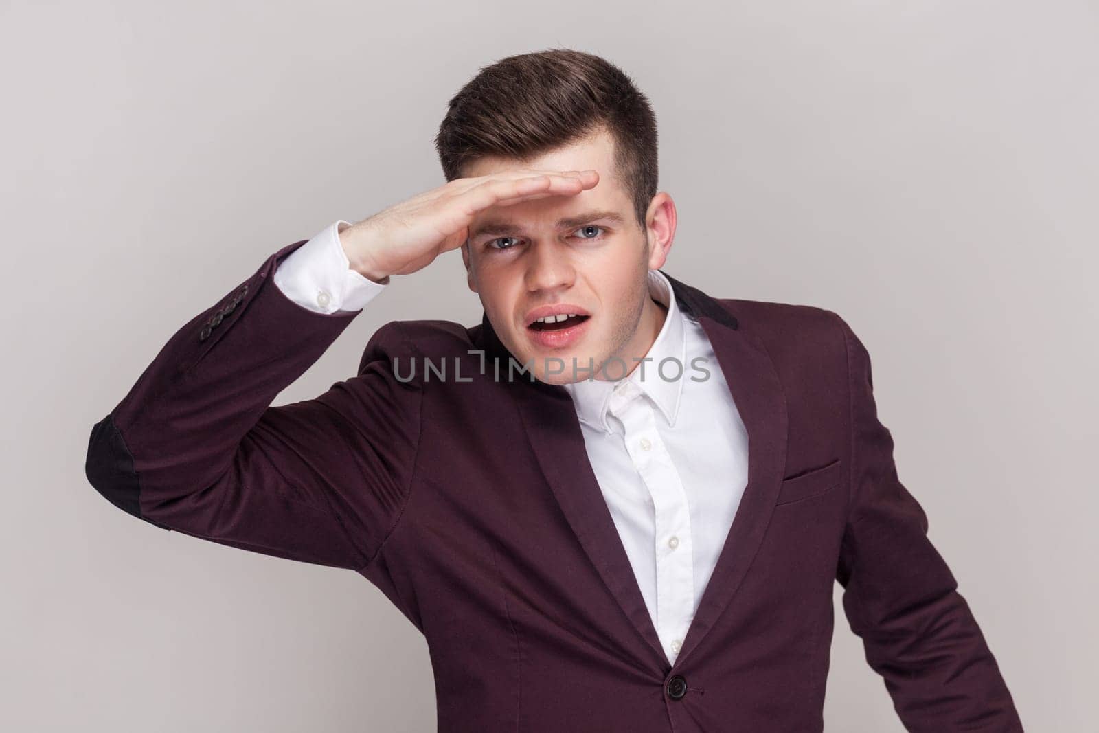 Man peering into distance, looking far away with attentive view, searching on horizon and curious to discover, wearing violet suit and white shirt. Indoor studio shot isolated on grey background.