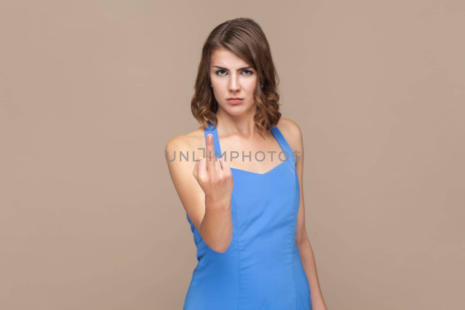 Portrait of displeased woman with wavy hair showing fuck sign, showing her negative feeling, looking at camera, wearing blue dress. Indoor studio shot isolated on light brown background.