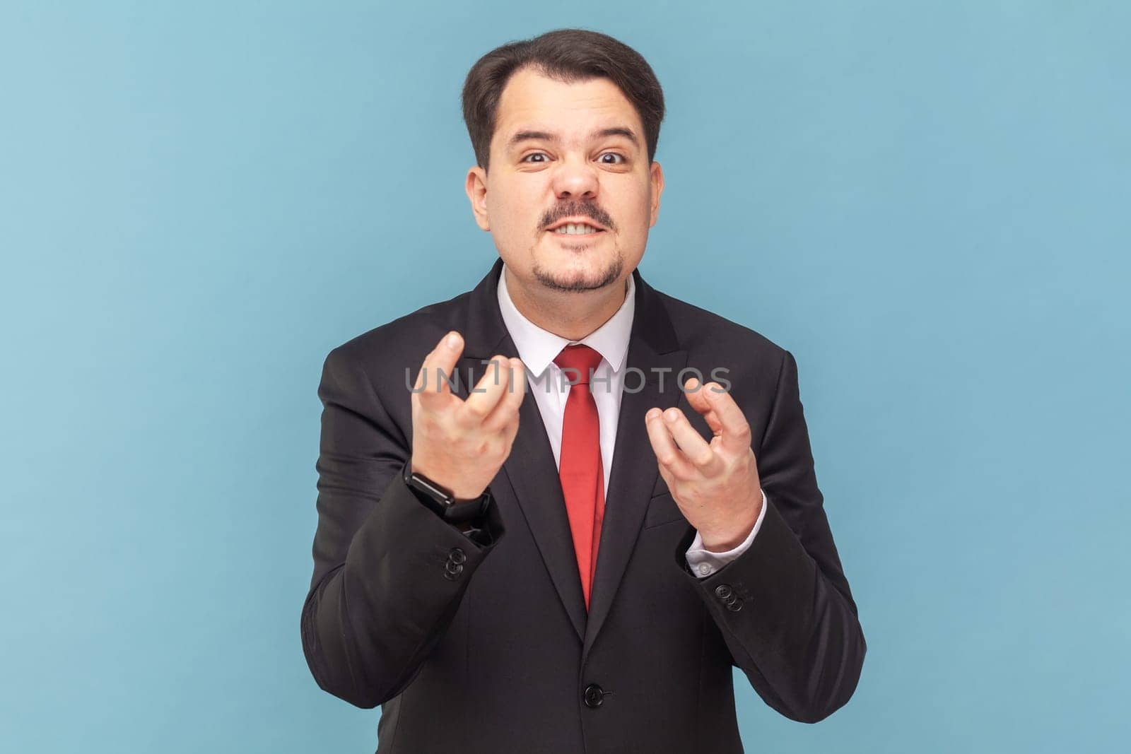 Portrait of angry aggressive man with mustache standing with raised arms, expressing anger and hate, wearing black suit with red tie. Indoor studio shot isolated on light blue background.