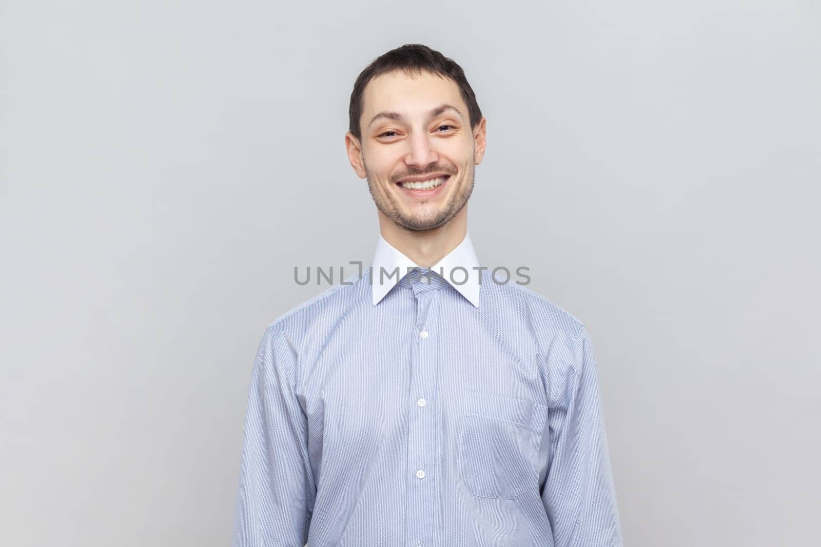 Portrait of handsome smiling joyful man standing looking at camera with toothy smile, expressing happiness, wearing light blue shirt. Indoor studio shot isolated on gray background.