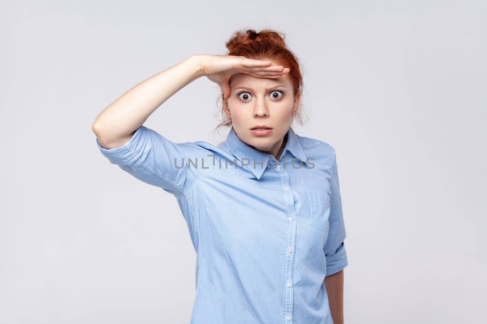 Portrait of scared ginger woman wearing blue shirt keeping palm over head and looking attentively far away, peering at long distance. Indoor studio shot isolated on gray background.
