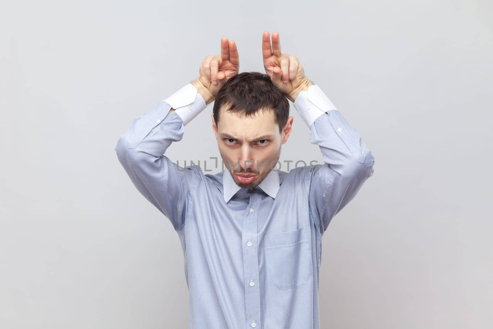 Portrait of angry serious aggressive man standing and showing bully horns, attacks, looks crazy and dangerous, wearing light blue shirt. Indoor studio shot isolated on gray background.