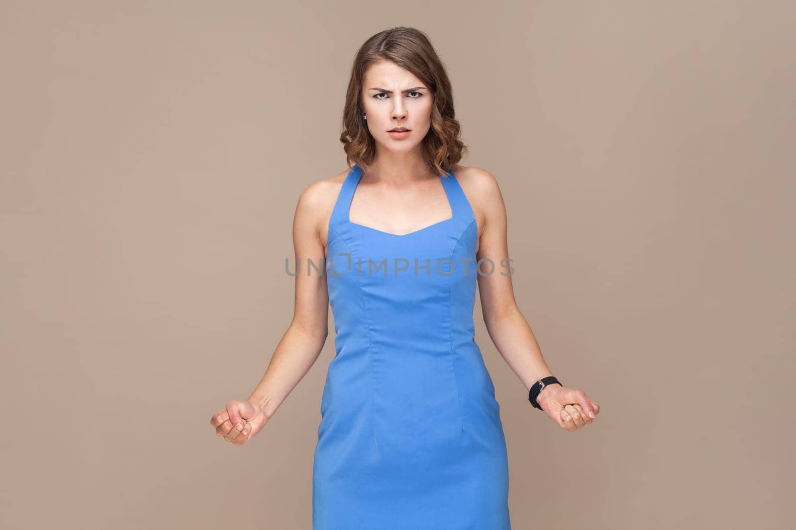 Angry aggressive young adult woman with wavy hair standing with clenched fists, looking at camera with hate and anger, wearing blue dress. Indoor studio shot isolated on light brown background.