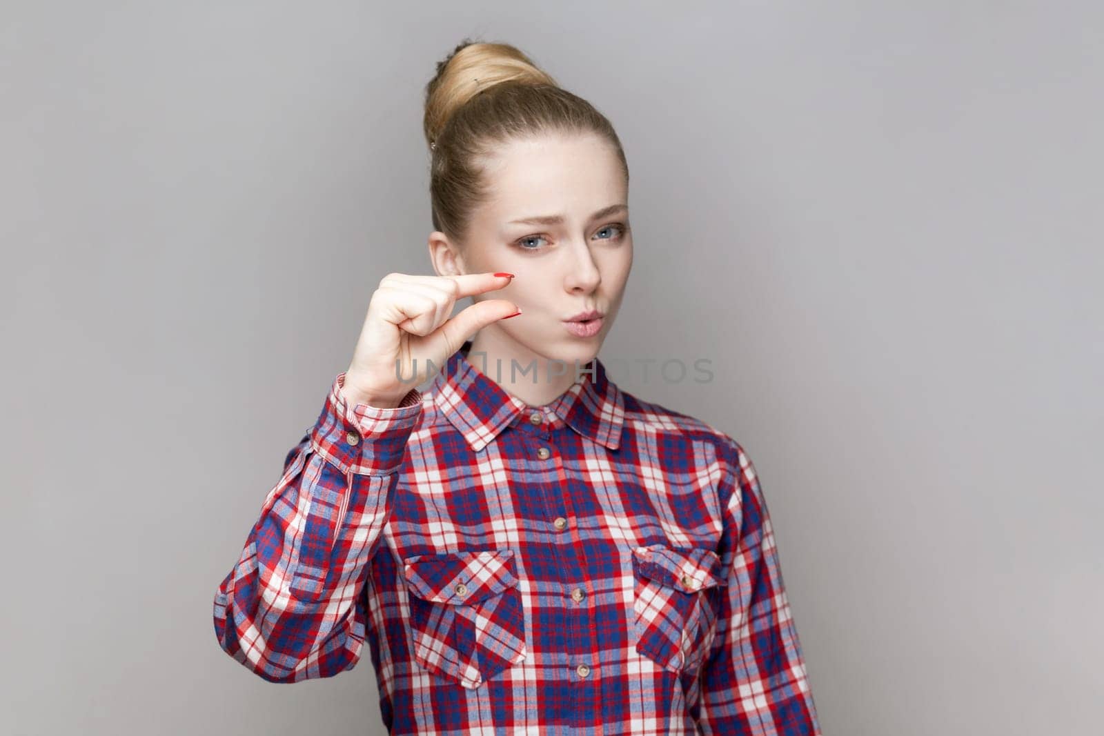 Portrait of beautiful adorable woman with bun hairstyle standing looking at camera, showing small gesture with fingers, wearing checkered shirt. Indoor studio shot isolated on gray background.