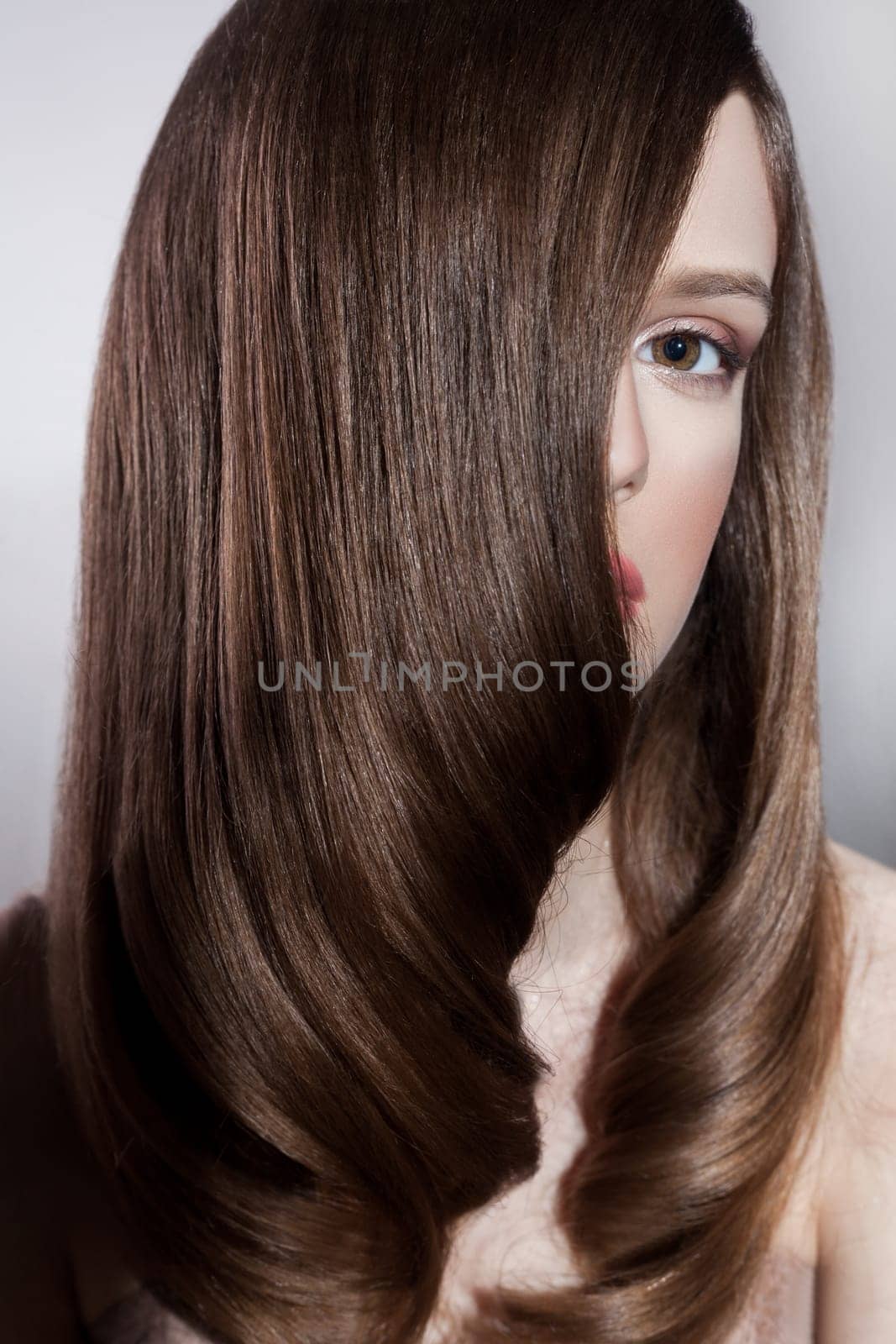 Portrait of attractive brunette woman fashion model with silk straight shiny hair and makeup, covering half of face with hair, looking at camera. Indoor studio shot isolated on gray background.