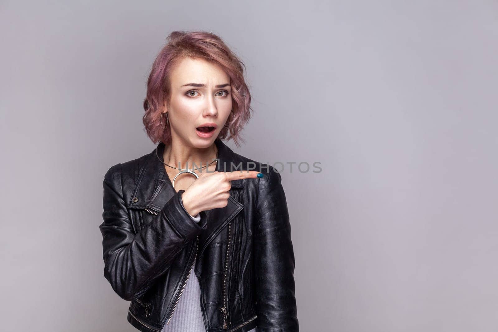 Portrait of shocked surprised woman with short hairstyle standing pointing away at copy space for advertisement, wearing black leather jacket. Indoor studio shot isolated on grey background.