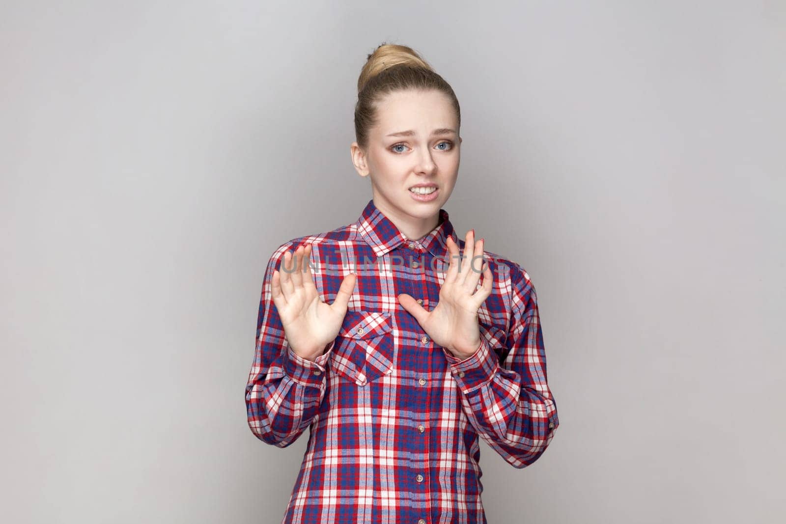 Portrait of scared frighten beautiful woman with bun hairstyle standing looking at camera, showing stop signs, being afraid, wearing checkered shirt. Indoor studio shot isolated on gray background.