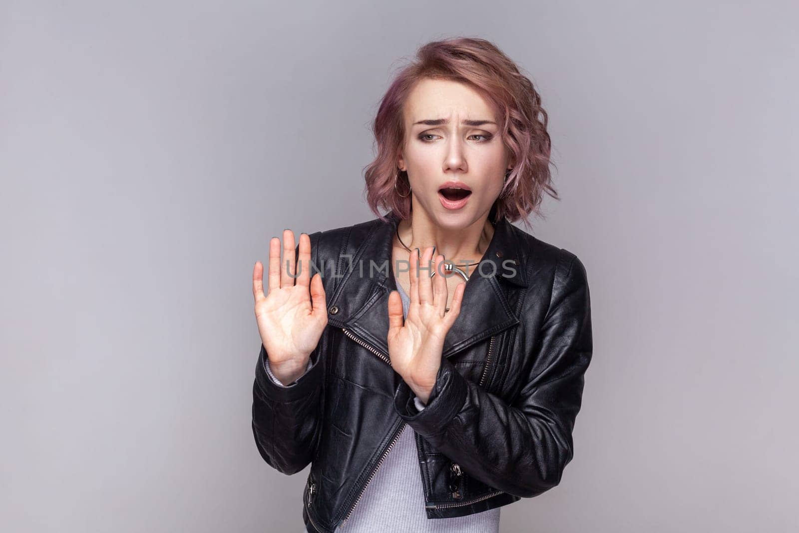 Portrait of scared frighten shocked woman with short hairstyle standing with showing stop sign, asking to stop, wearing black leather jacket. Indoor studio shot isolated on grey background.