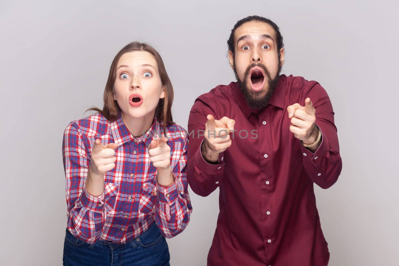 Portrait of shocked amazed woman and man standing together and pointing at camera, choosing you, looking at camera with big eyes. Indoor studio shot isolated on gray background.