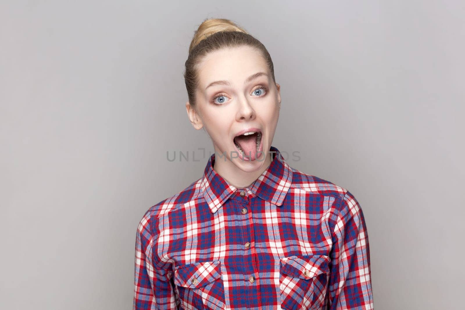 Portrait of foolish crazy playful woman with bun hairstyle looking at camera with open mouth and showing tongue out, wearing checkered shirt. Indoor studio shot isolated on gray background.