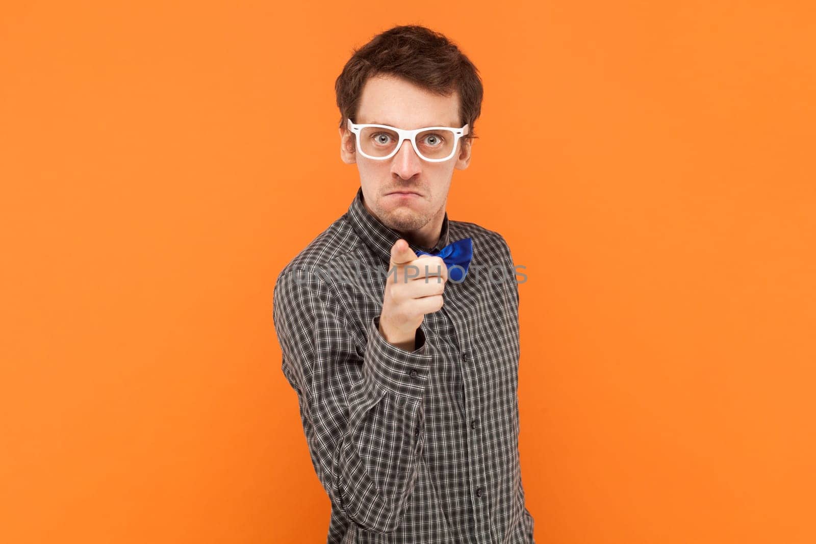 Self assured serious man nerd points index finger at camera chooses you looks directly at camera, wearing shirt with blue bow tie and white glasses. Indoor studio shot isolated on orange background.