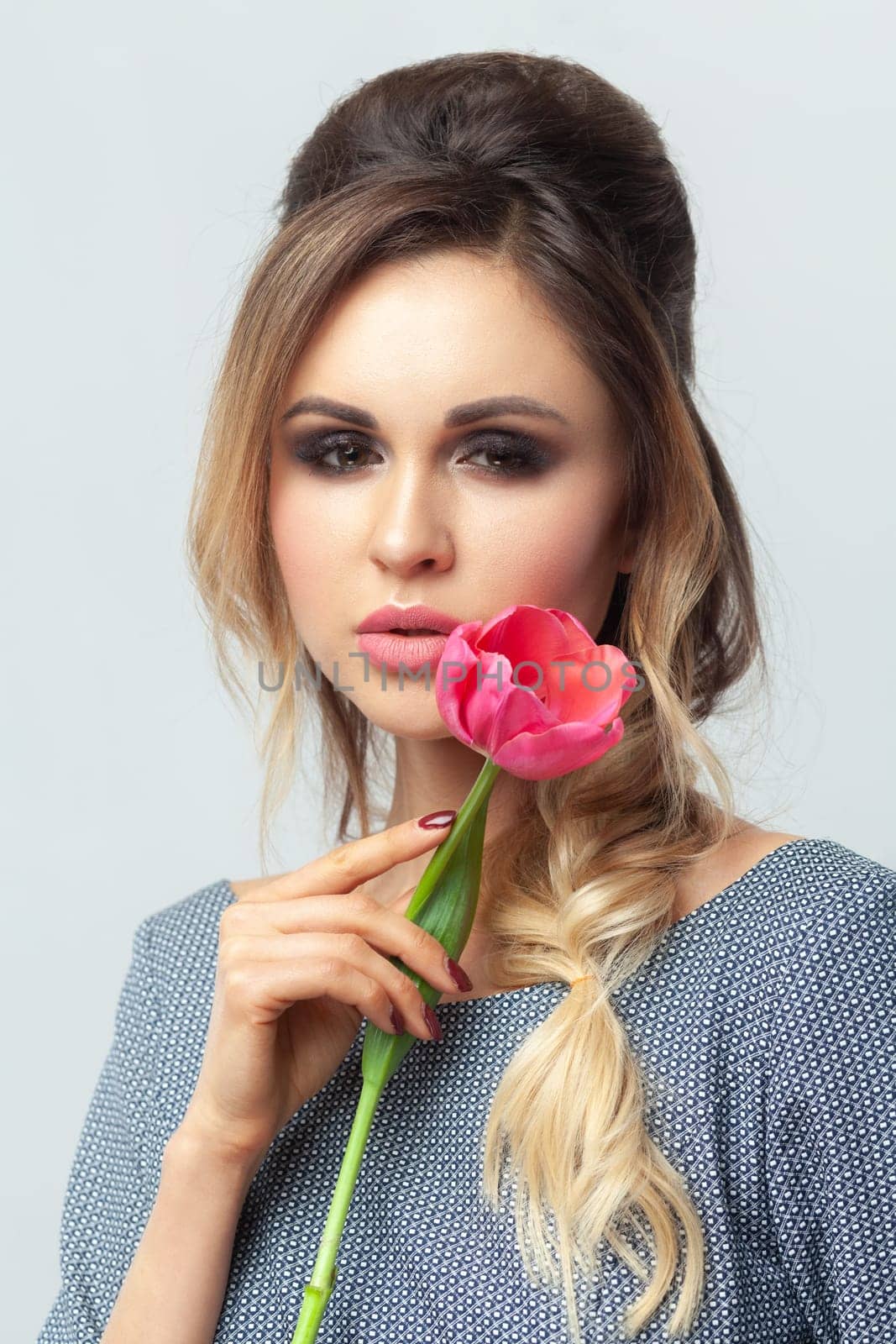 Closeup portrait of attractive gentle blonde woman fashion model with makeup and hairstyle, holding tulip near her face, wearing grey elegant dress. Indoor studio shot isolated on gray background.