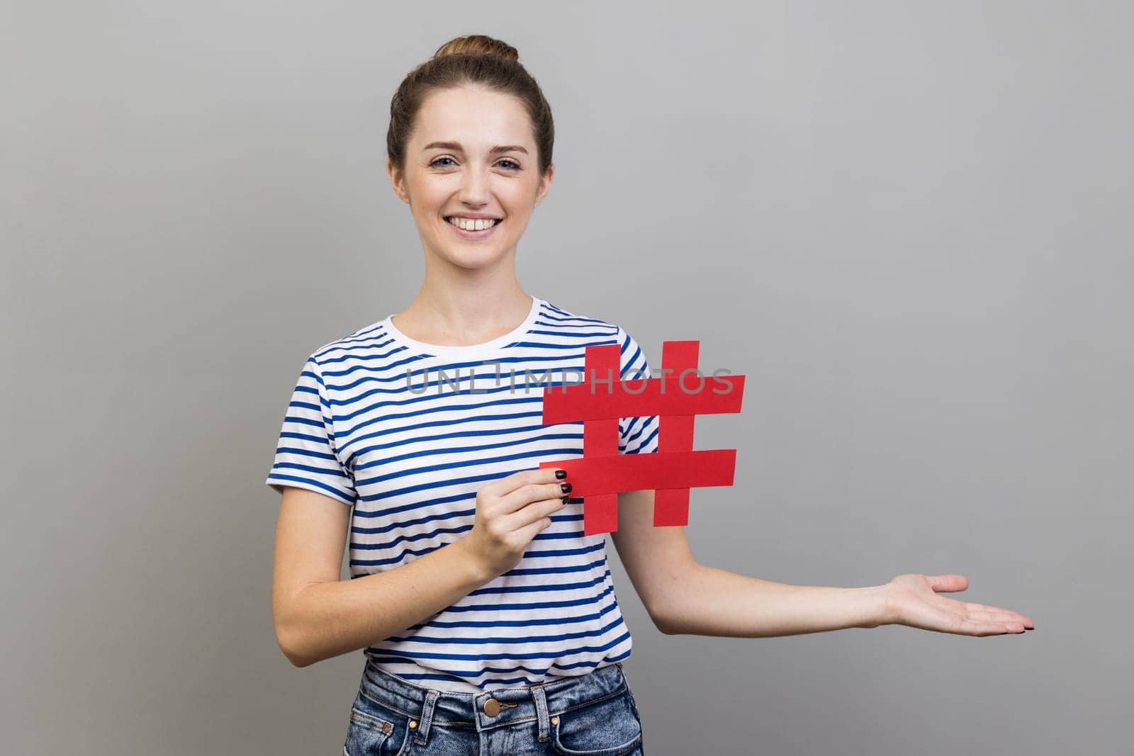 Portrait of woman in striped T-shirt holding hashtag and presenting at copy space on her palm, empty place for idea presentation, product advertising. Indoor studio shot isolated on gray background.