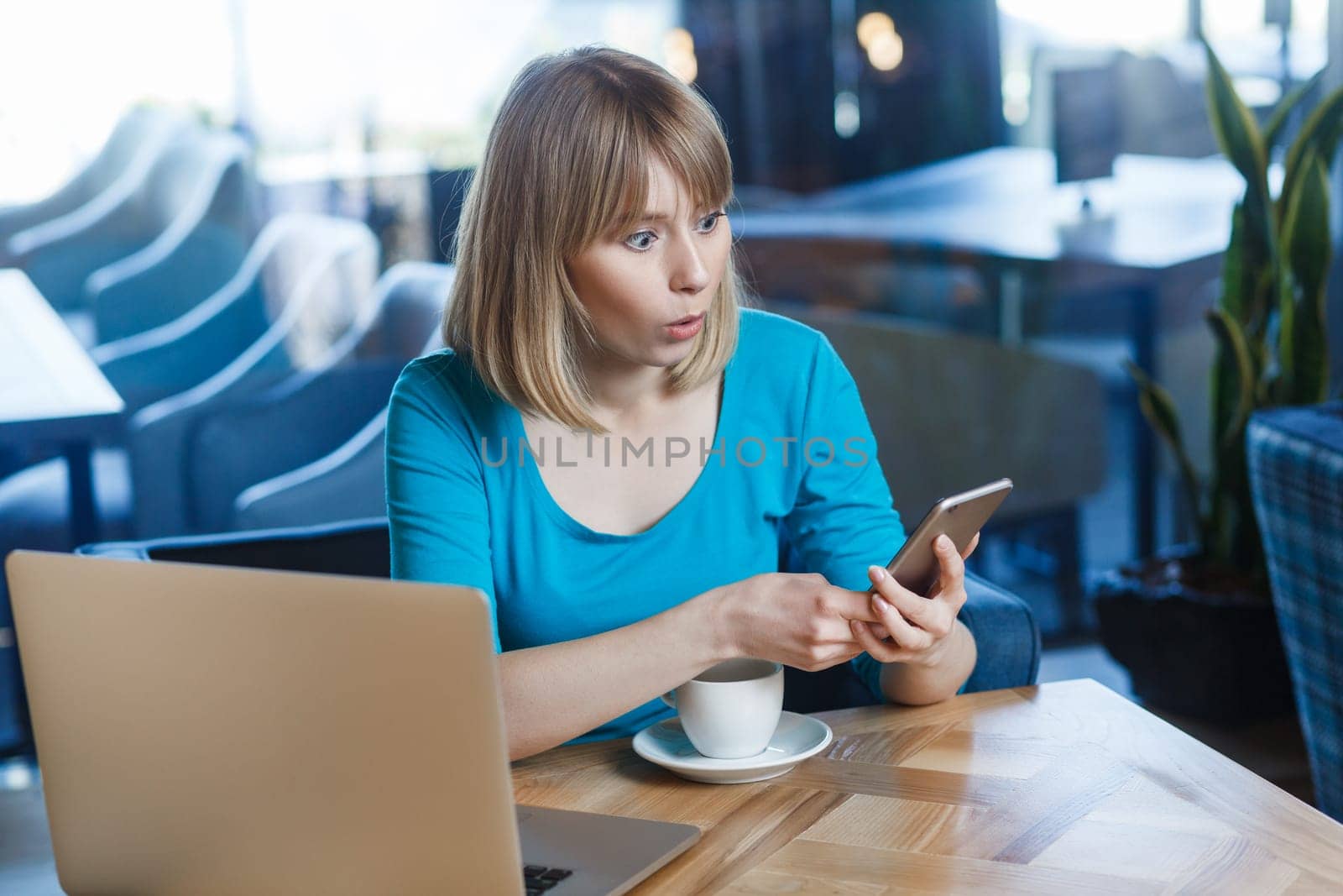 Portrait of shocked amazed young woman with blonde hair in blue shirt working on laptop, having break, using cell phone, checking social networks, reading breaking news. Indoor shot in cafe.