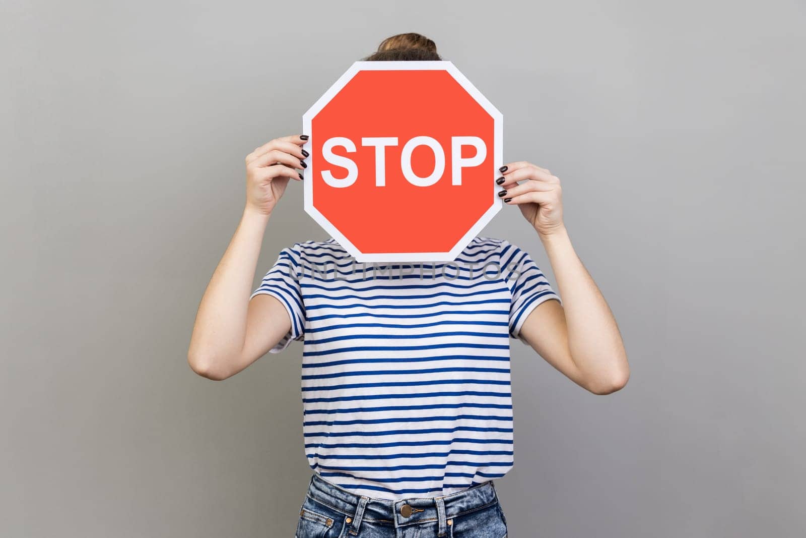 Portrait of woman wearing striped T-shirt hiding her face behind stop sign, afraid talking about domestic violence, scared victim. Indoor studio shot isolated on gray background.