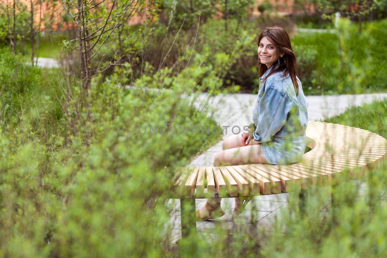 Beautiful woman sitting on wooden bench in green park, looking at camera with toothy smile, by Khosro1