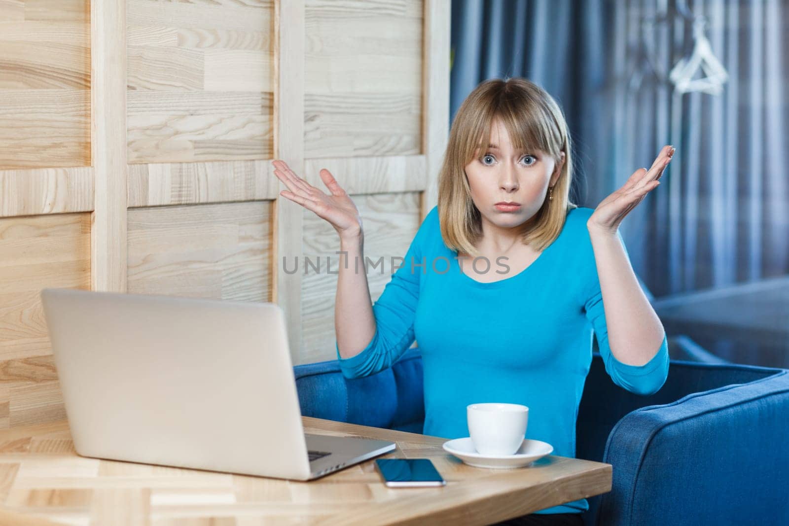 Confused uncertain young woman with blonde hair in blue shirt sitting at table and working on laptop, shrugging shoulders, looking at camera with puzzled face. Indoor shot, cafe background.