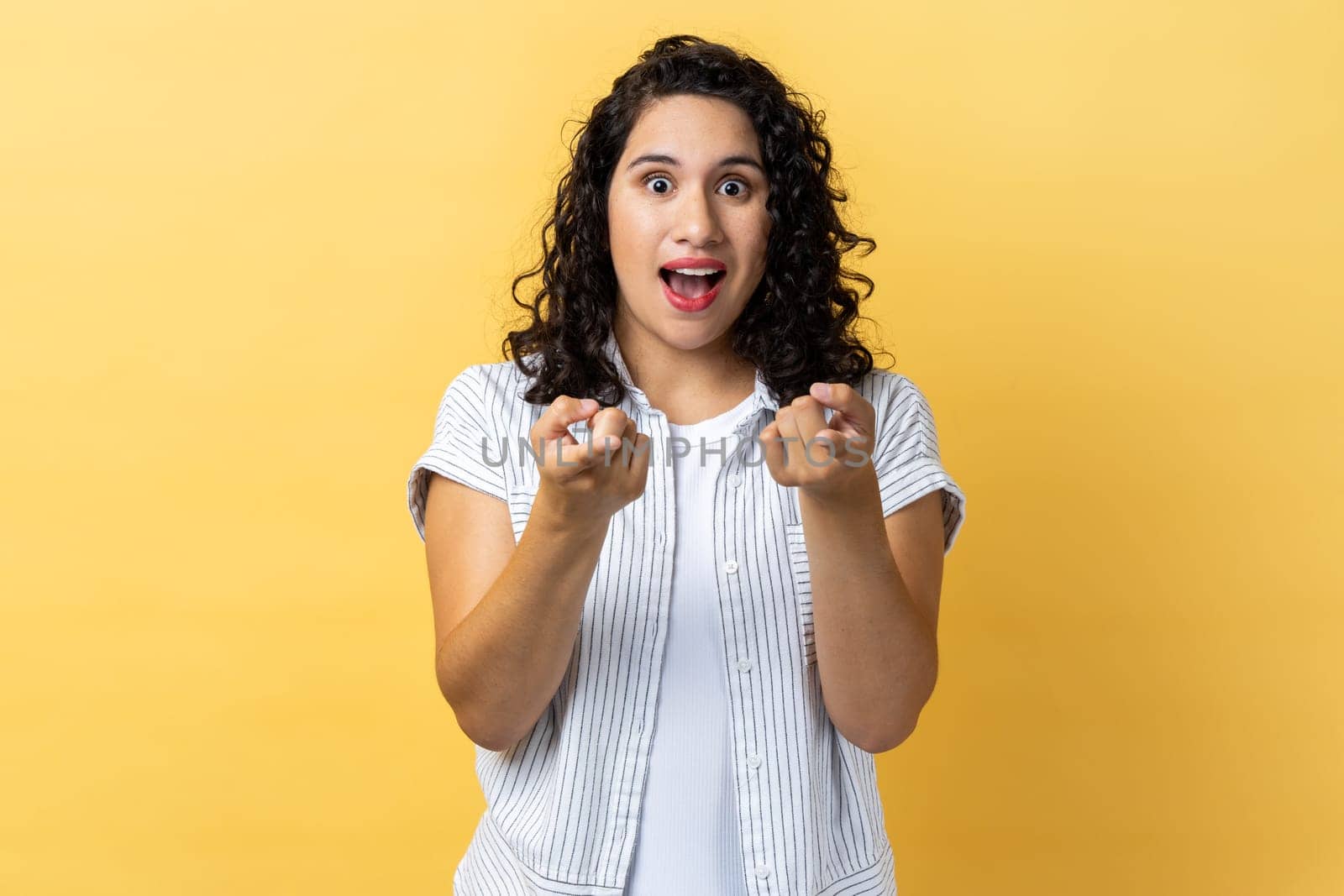 Portrait of amazed excited beautiful woman with dark wavy hair pointing directly at you, choosing something, has shocked expression. Indoor studio shot isolated on yellow background.