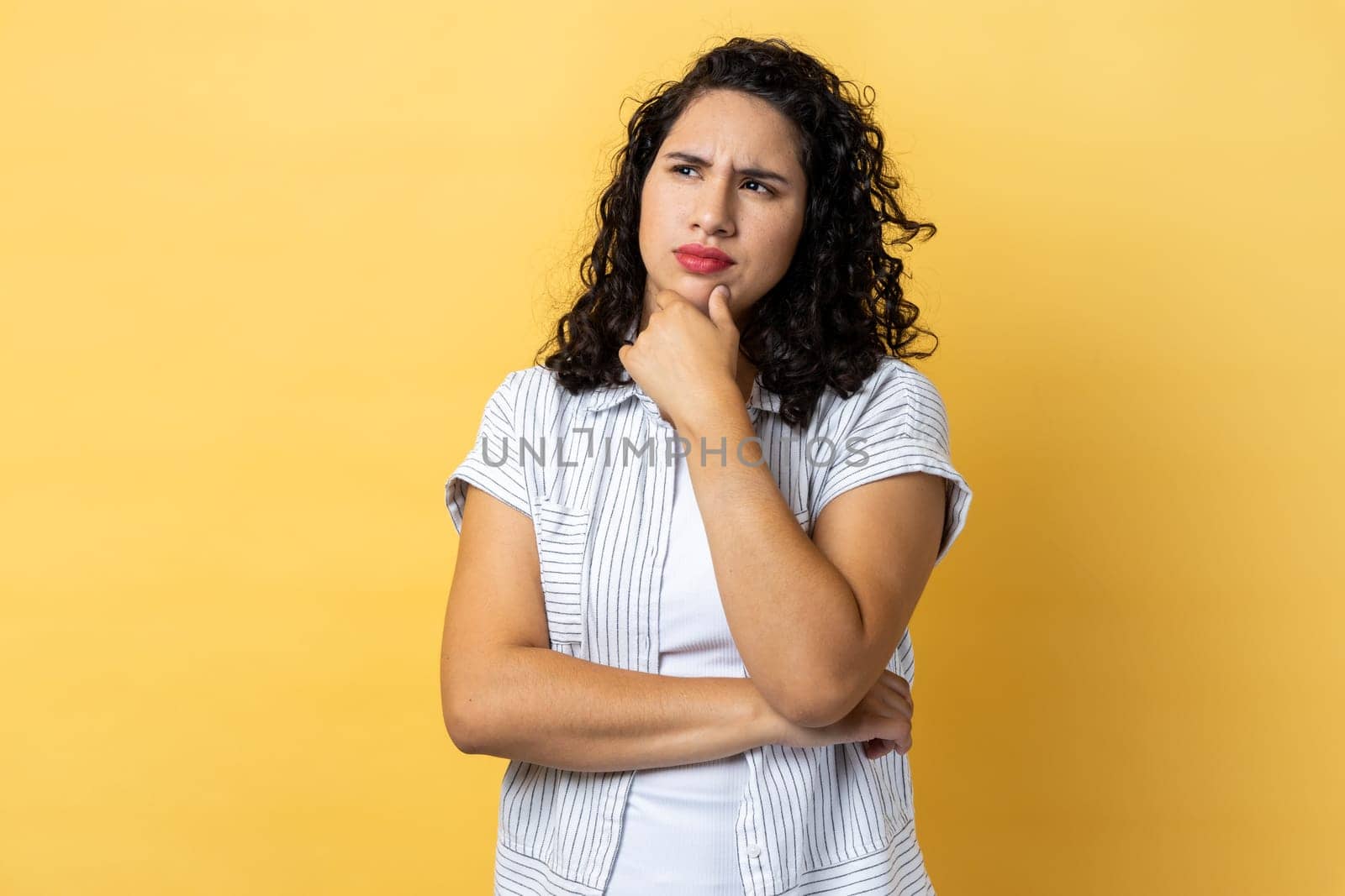 Portrait of pensive attractive woman with dark wavy hair being deeply in thoughts, keeping hand on head, looking away, holding chin. Indoor studio shot isolated on yellow background.