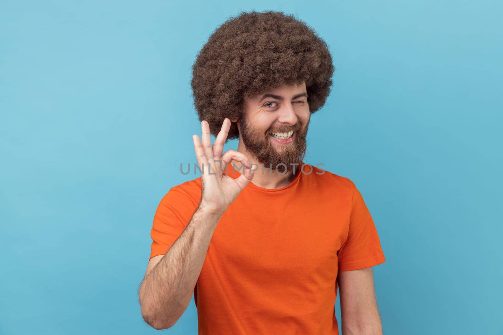 Portrait of positive man with Afro hairstyle wearing orange T-shirt standing and looking at camera with Ok sign, winking and toothy smile. Indoor studio shot isolated on blue background.