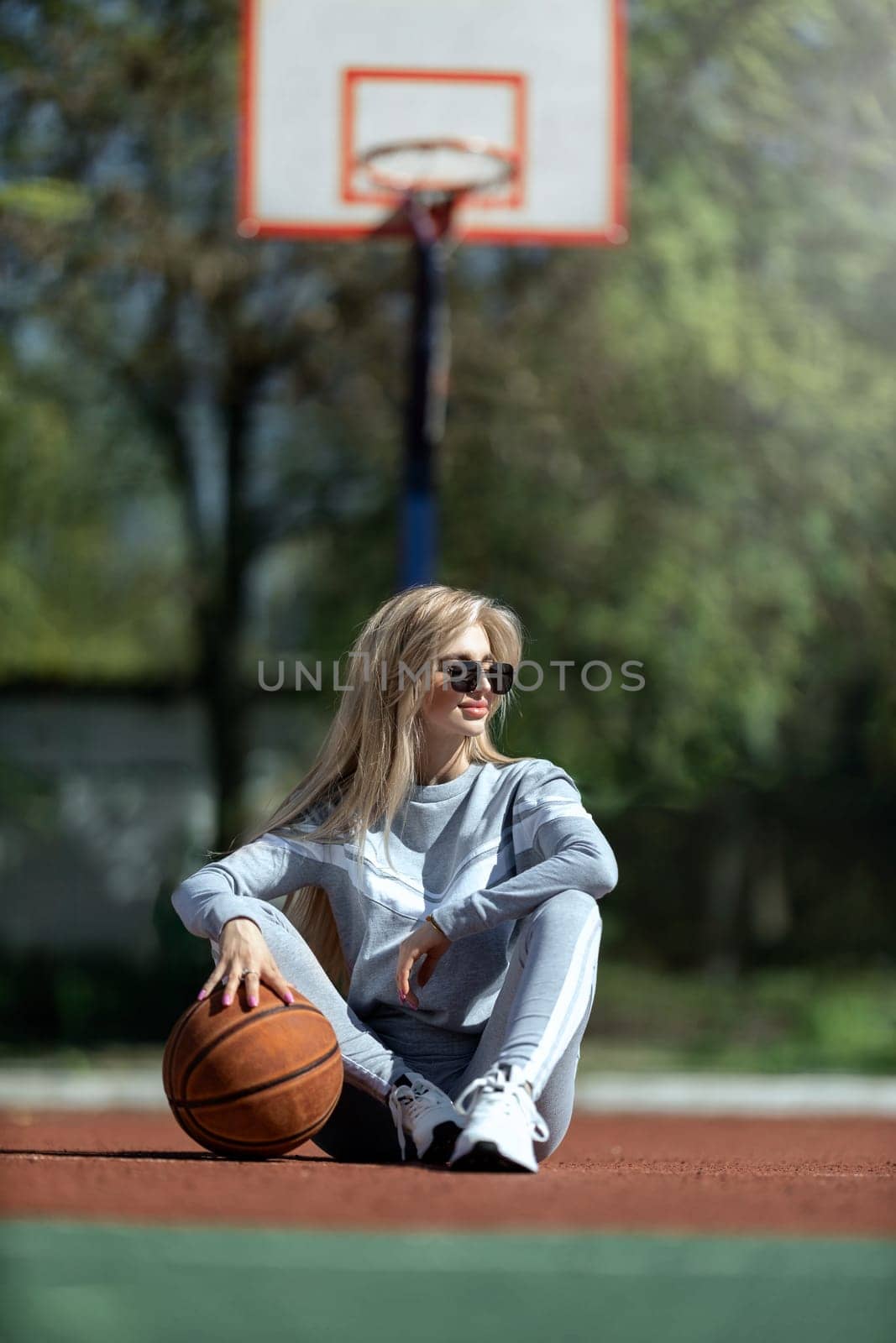 athletic blonde girl sits on the basketball court in a gray tracksuit with a ball in her hands