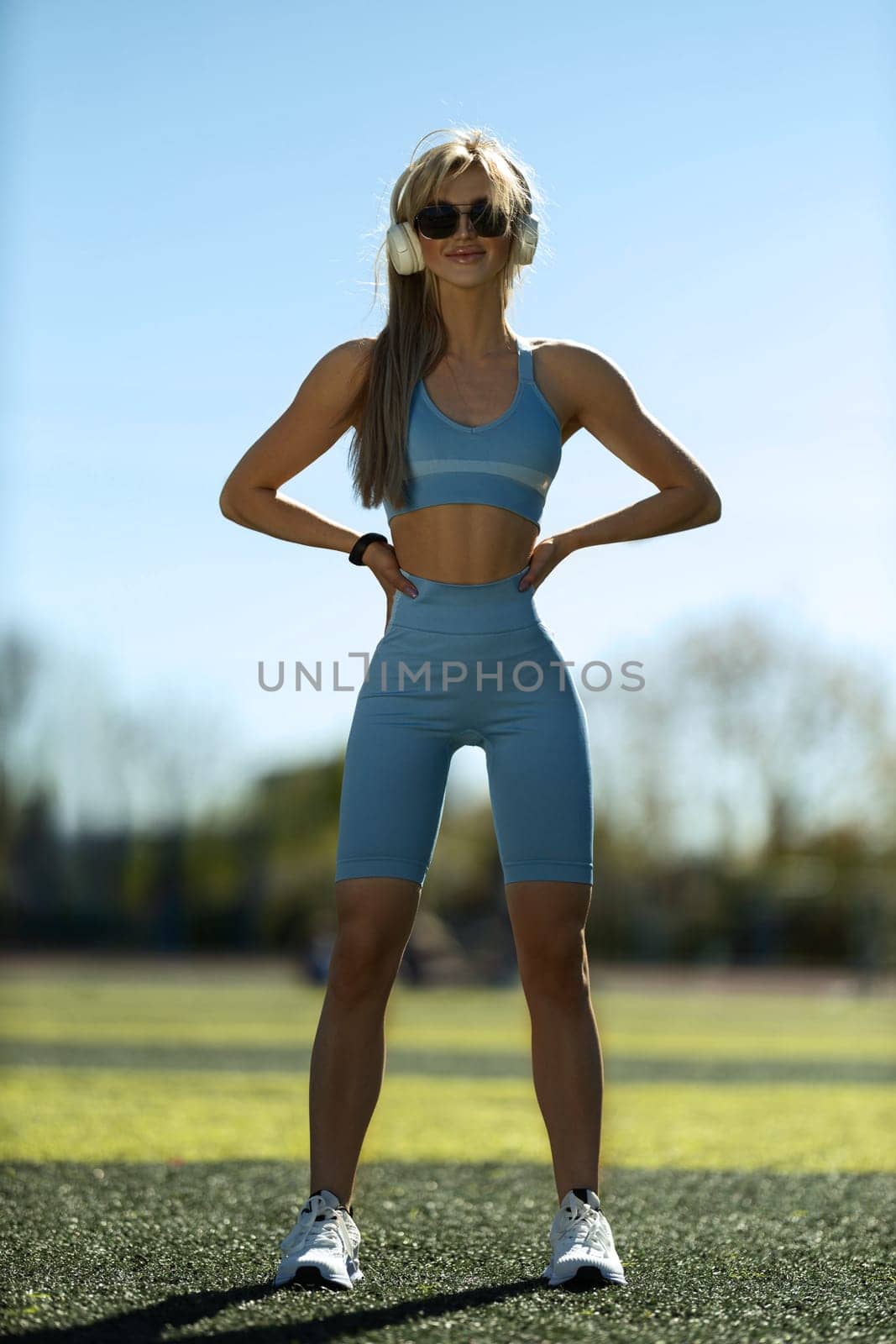 blonde athletic girl stands on the football field in a tank top, breeches, white sneakers and white headphones