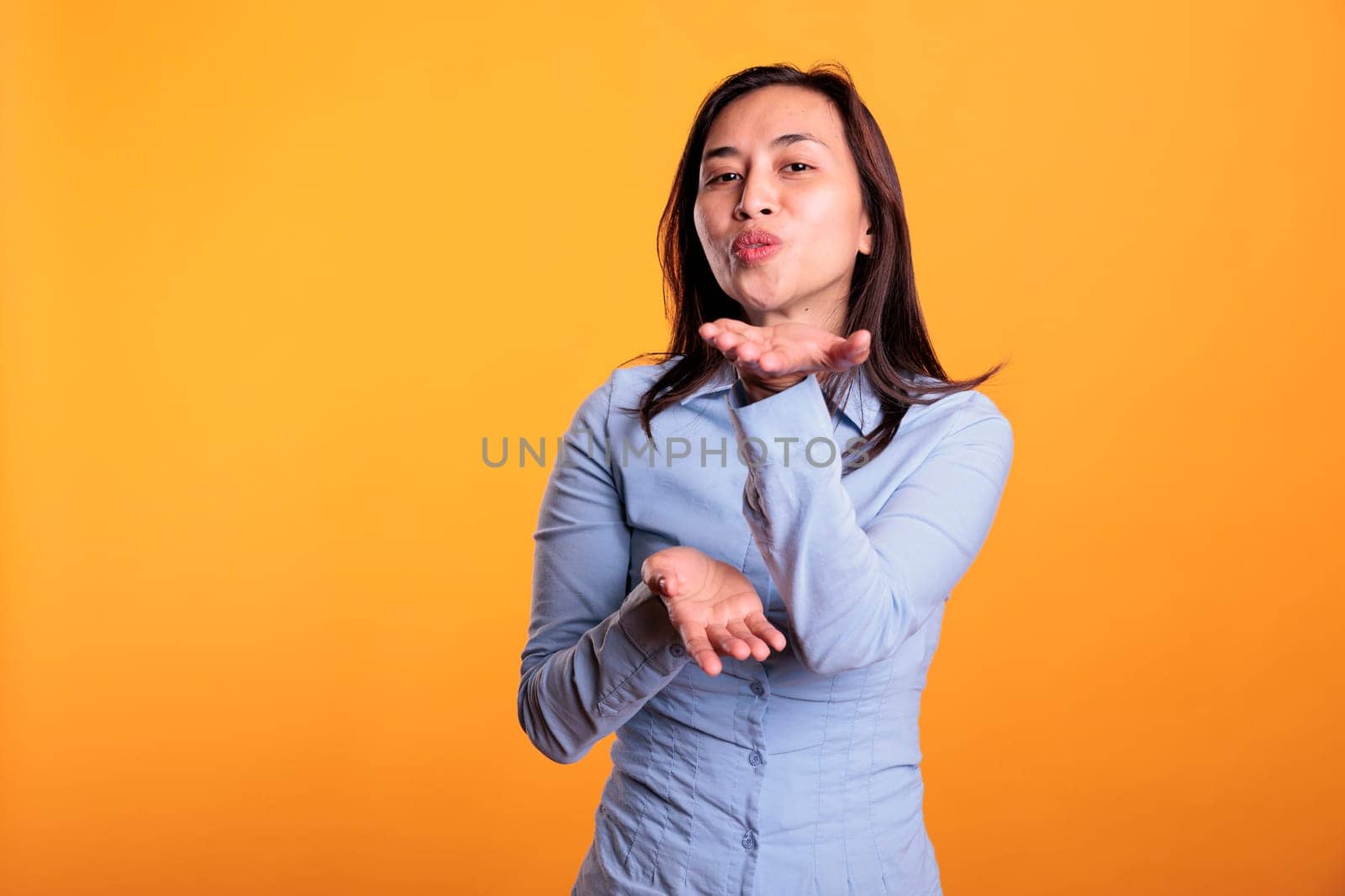 Confident joyful filipino woman blowing air kissed during studio shot, posing over yellow background. Attractive model expressing love, looking at the camera with a flirty smile. Romantic gesture