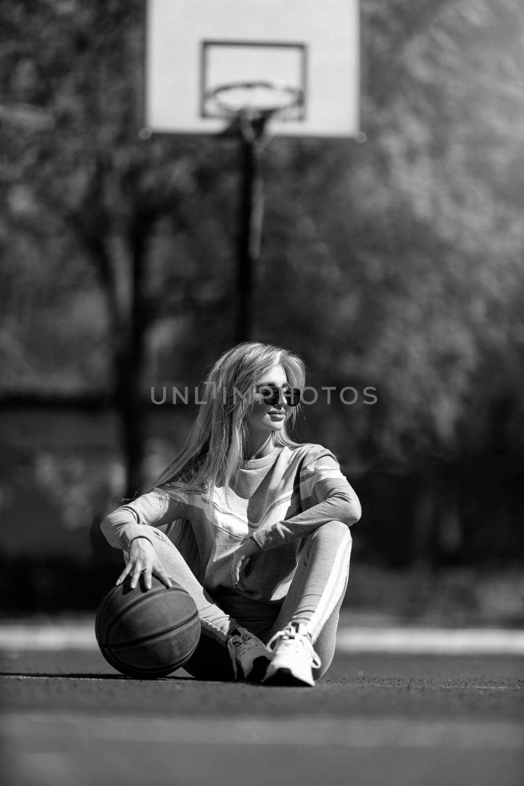 athletic blonde girl sits on the basketball court in a gray tracksuit with a ball in her hands black and white