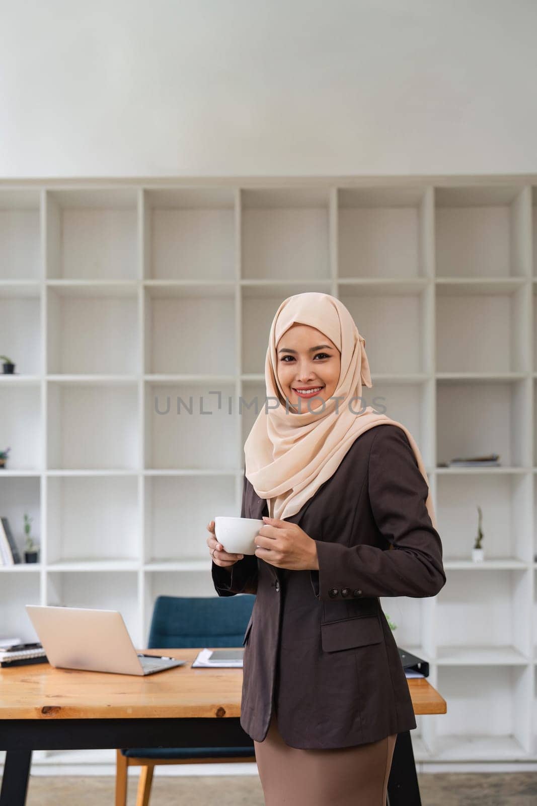Asian Muslim business woman holding a coffee mug standing in the modern office. business people, diversity and office concept.