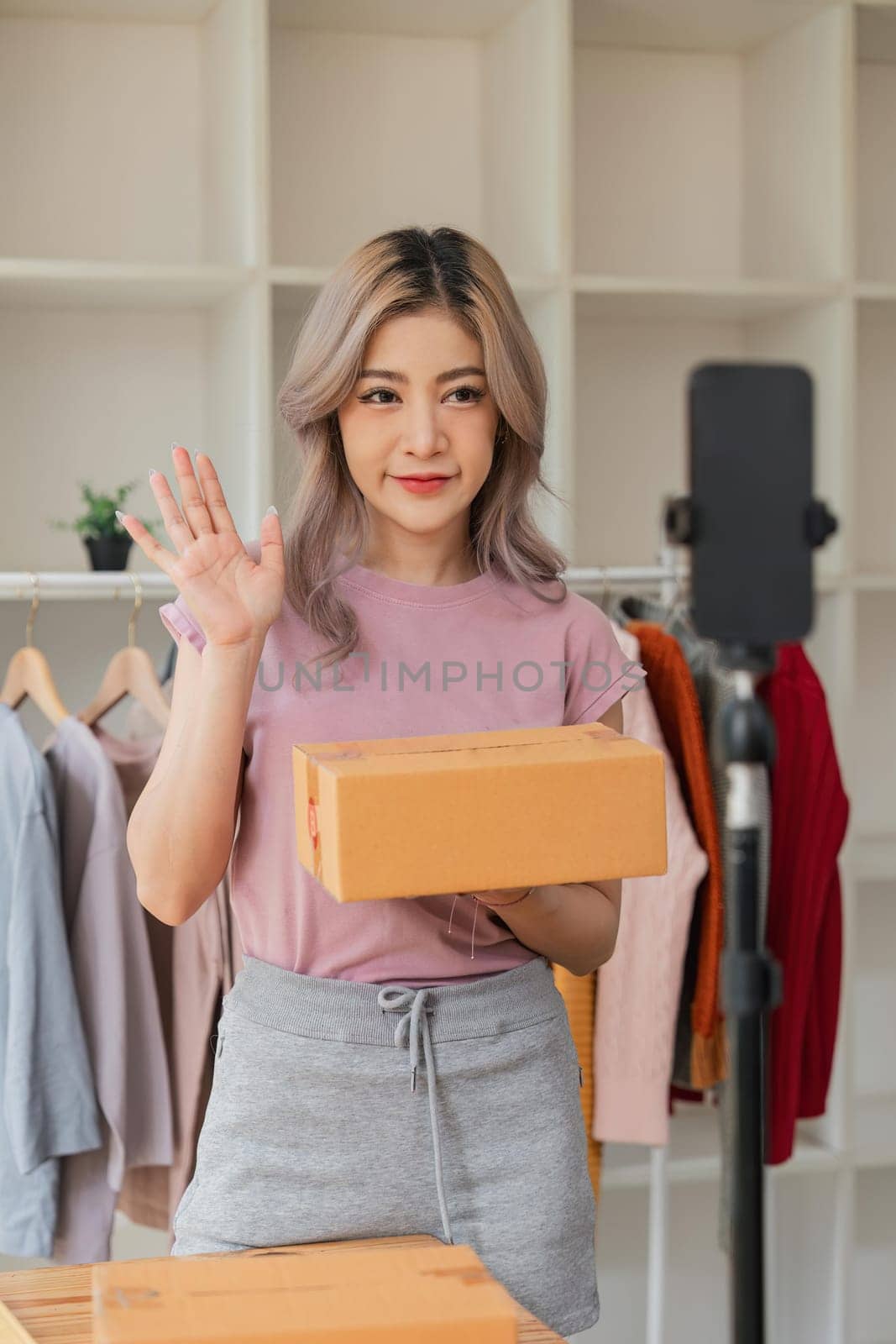 Woman entrepreneur showing clothes product on live online social media streaming and packing into cardboard box to preparing for shipping customer delivery at home.