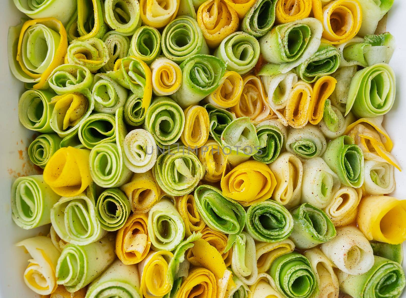 Small rolls of zucchini green and yellow with creamy sauce in a white baking dish against the background of a checkered napkin. Top view, close-up