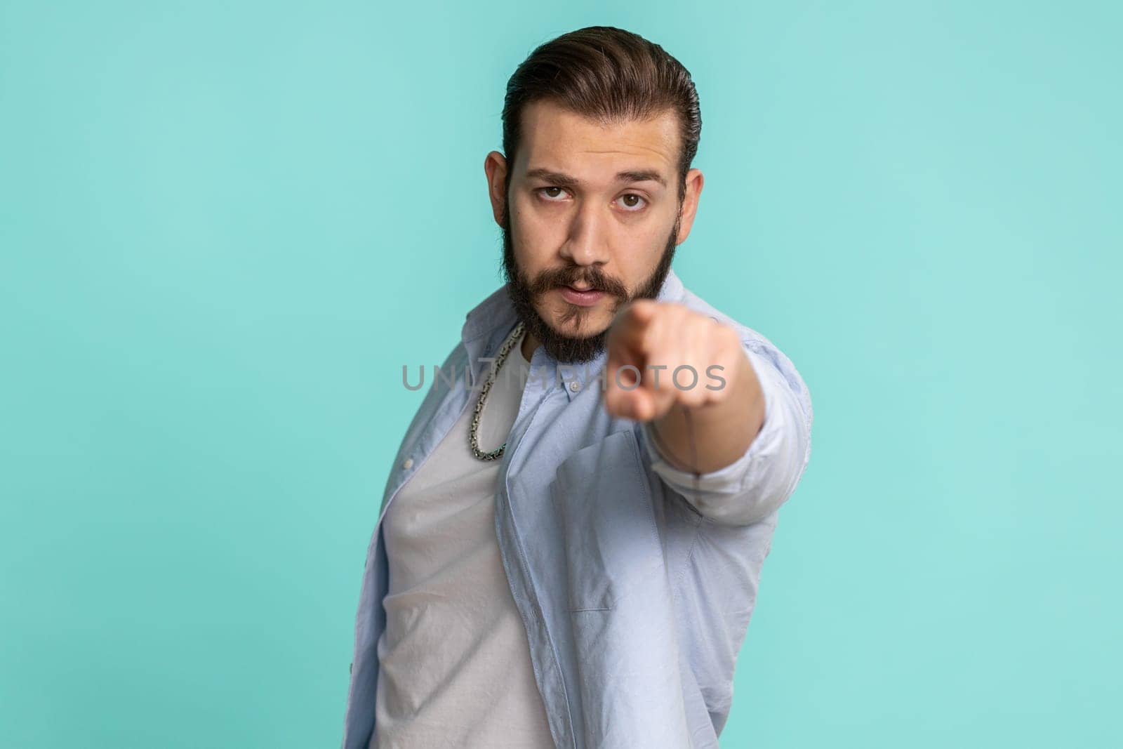 Hey you. Young lebanese man smiling excitedly and pointing to camera, choosing lucky happy winner, indicating to awesome you. Handsome bearded arabian guy isolated alone on blue studio wall background