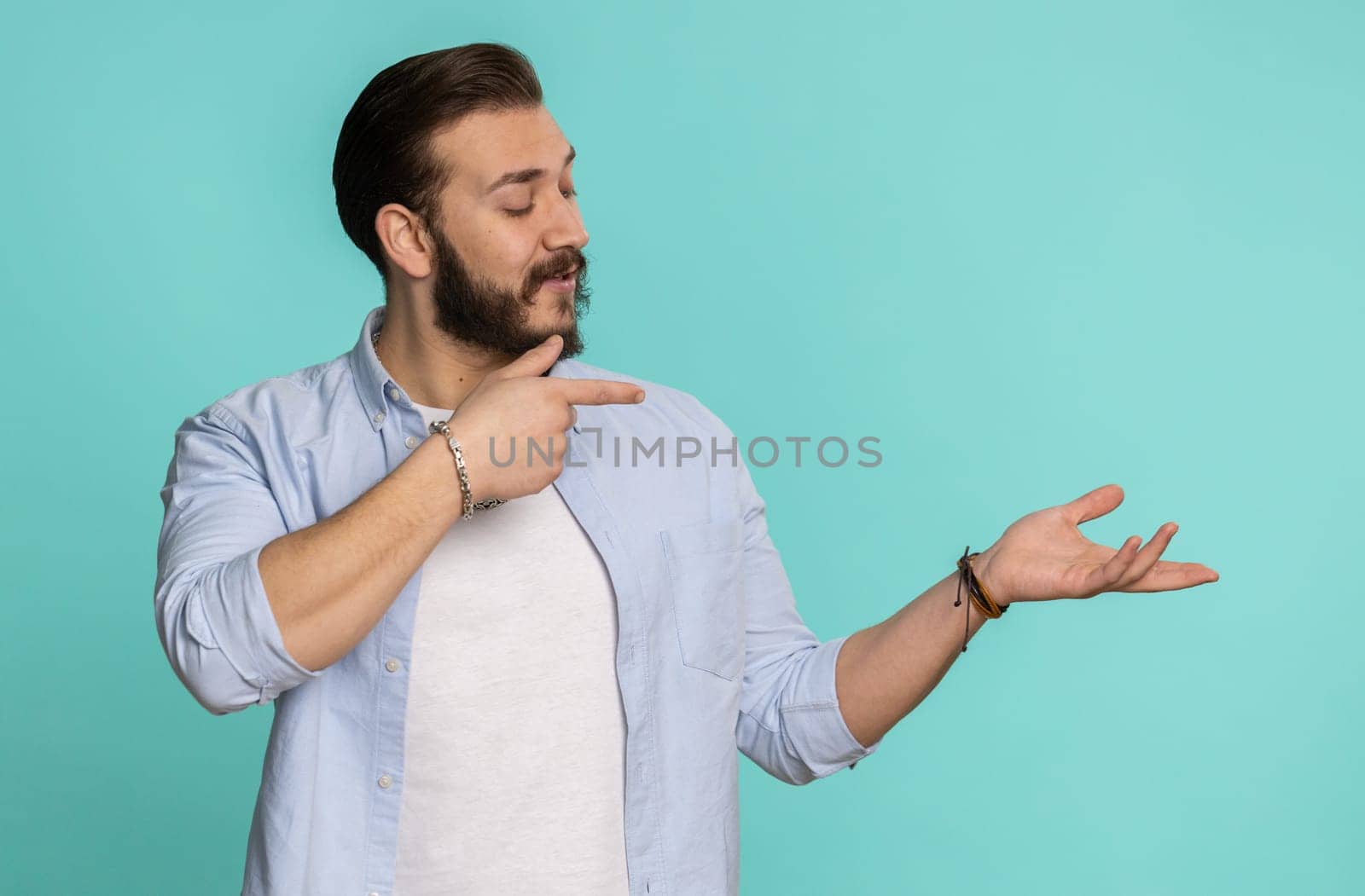 Young lebanese man showing thumbs up and pointing empty place, advertising area for commercial text, copy space for goods promotion. Bearded arabian guy isolated on blue studio background, indoors