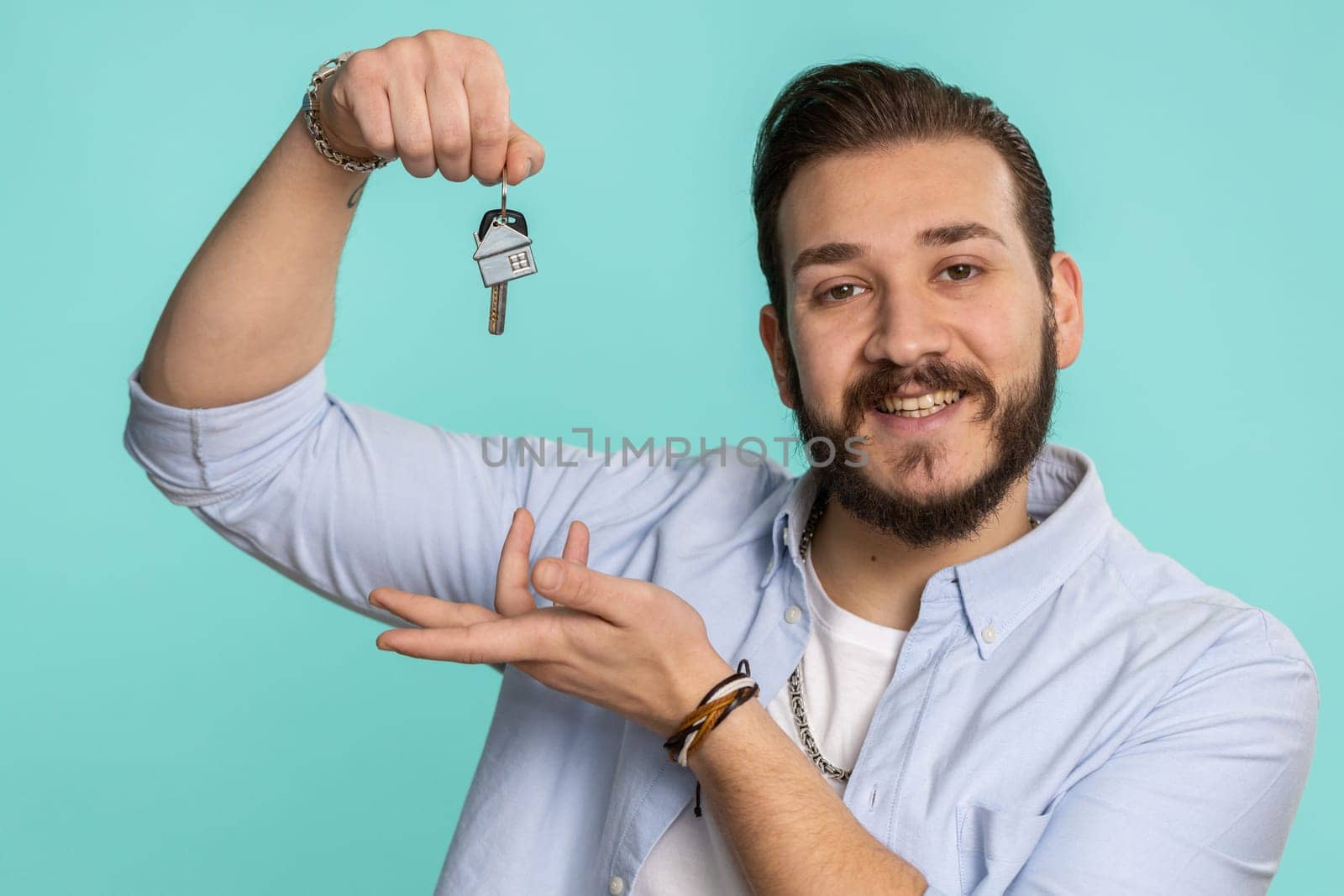 Young lebanese man real estate agent lifting hand showing the keys of new home house apartment, buying or renting property, mortgage loan. Bearded arabian guy isolated on blue studio background