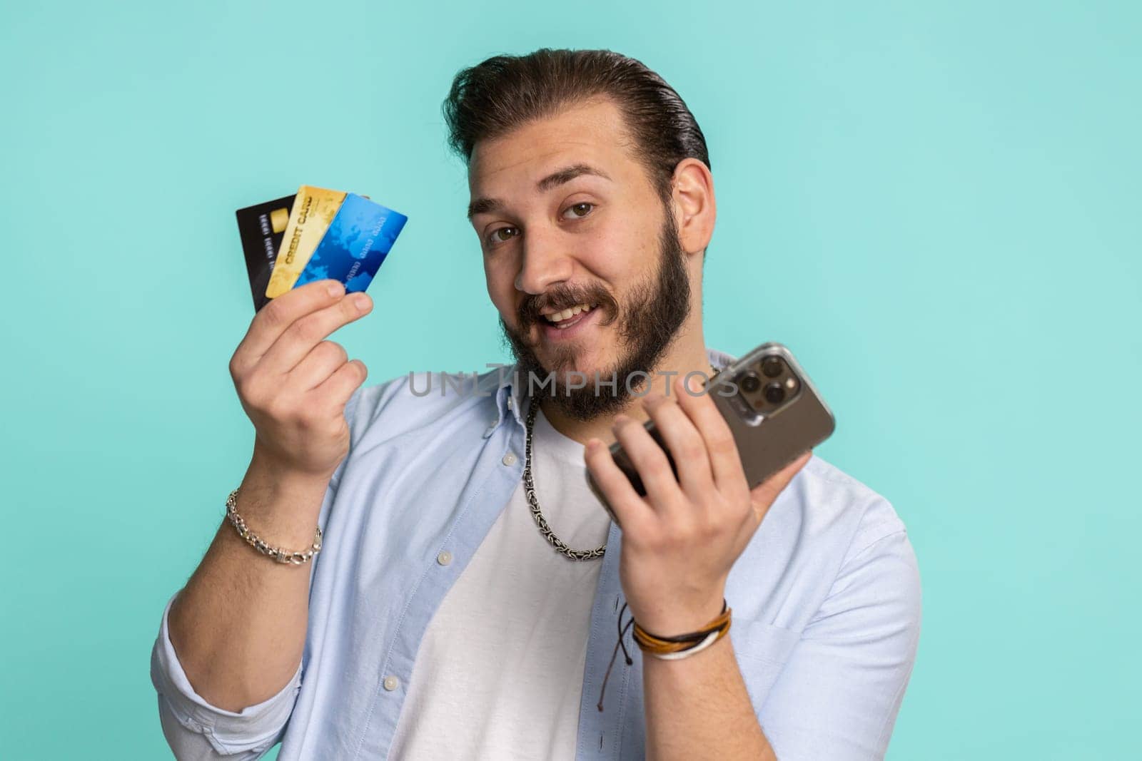 Sincere lebanese man customer using credit bank cards and smartphone while transferring money, purchases online shopping, payment. Finance and internet. Handsome guy isolated alone on blue background