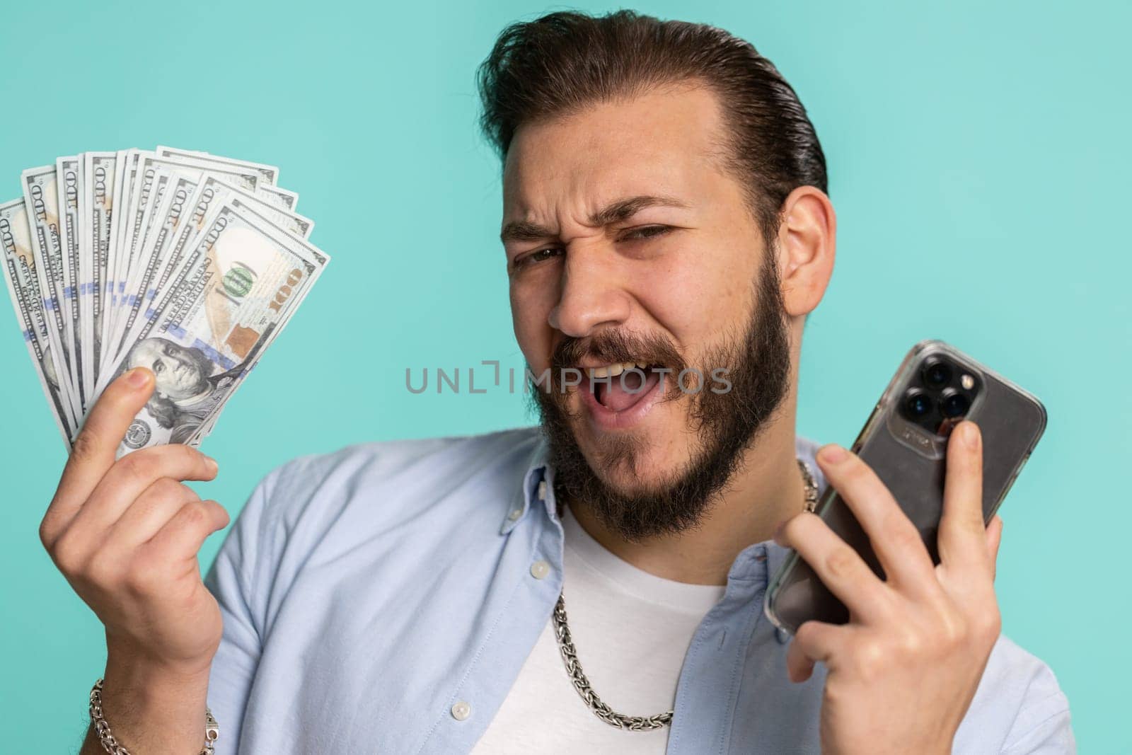 Lebanese man looking smartphone display sincerely rejoicing win, receiving money dollar cash banknotes, success lottery jackpot luck, income salary wage. Arabian guy isolated on studio blue background