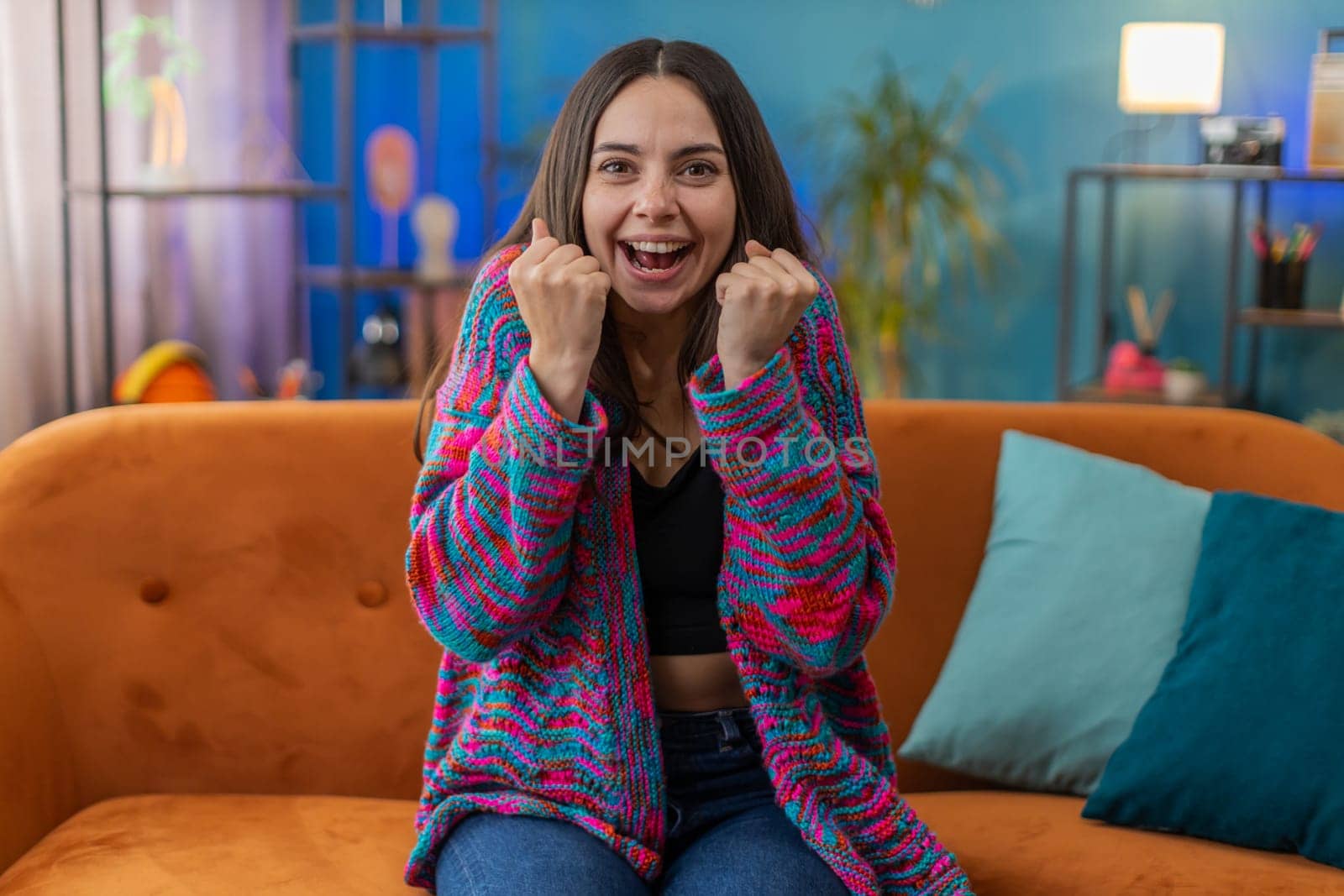 Happy Caucasian woman shouting, celebrating success, winning, goal achievement good news, lottery luck, victory, watching TV championship at modern home apartment. Girl in living room sitting on couch