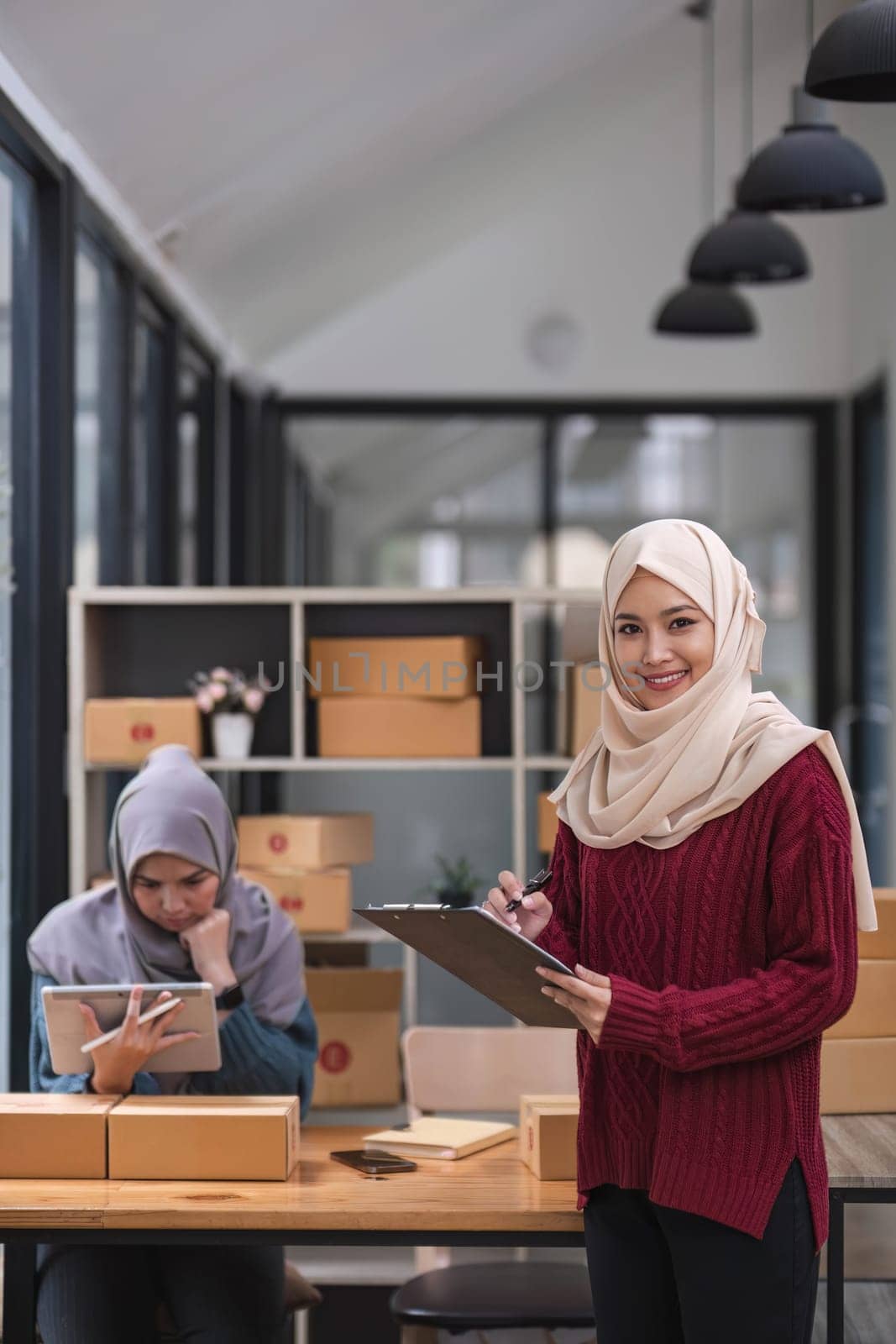 A happy young Asian Muslim female online seller working with her employee in the office. SME business, online shop owner by wichayada