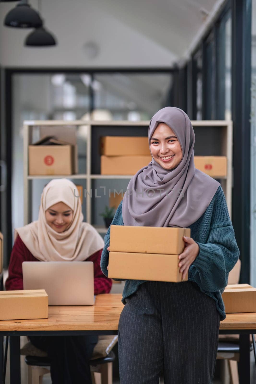 A happy young Asian Muslim female online seller working with her employee in the office. SME business, online shop owner..