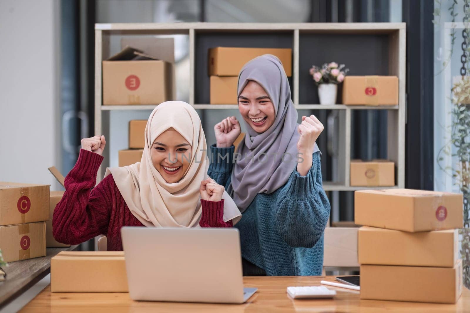 Two women muslim look at online product sales data on laptops and show joy by wichayada