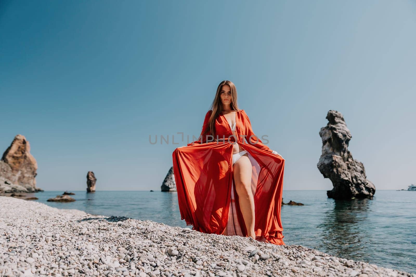 Woman travel sea. Happy tourist in red dress enjoy taking picture outdoors for memories. Woman traveler posing on the rock at sea bay surrounded by volcanic mountains, sharing travel adventure journey by panophotograph