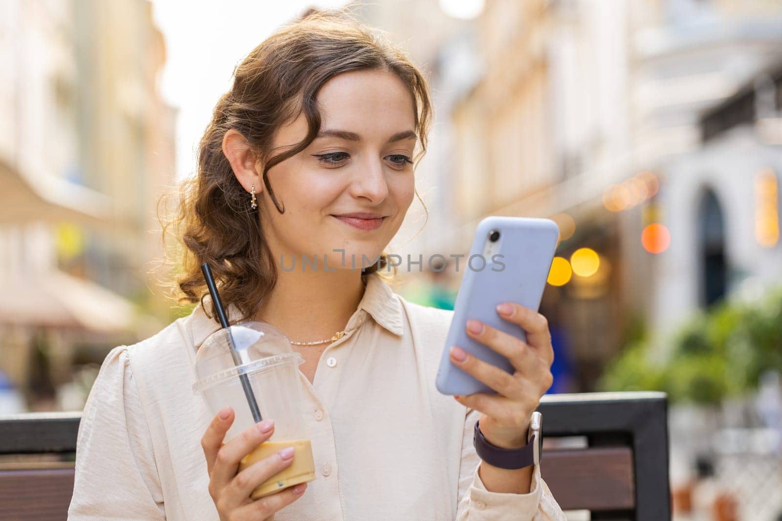 Smiling pretty young woman using smartphone typing text answering messages chatting online looking mobile screen social media app. Girl tourist sits on bench on city street drinks cold coffee with ice