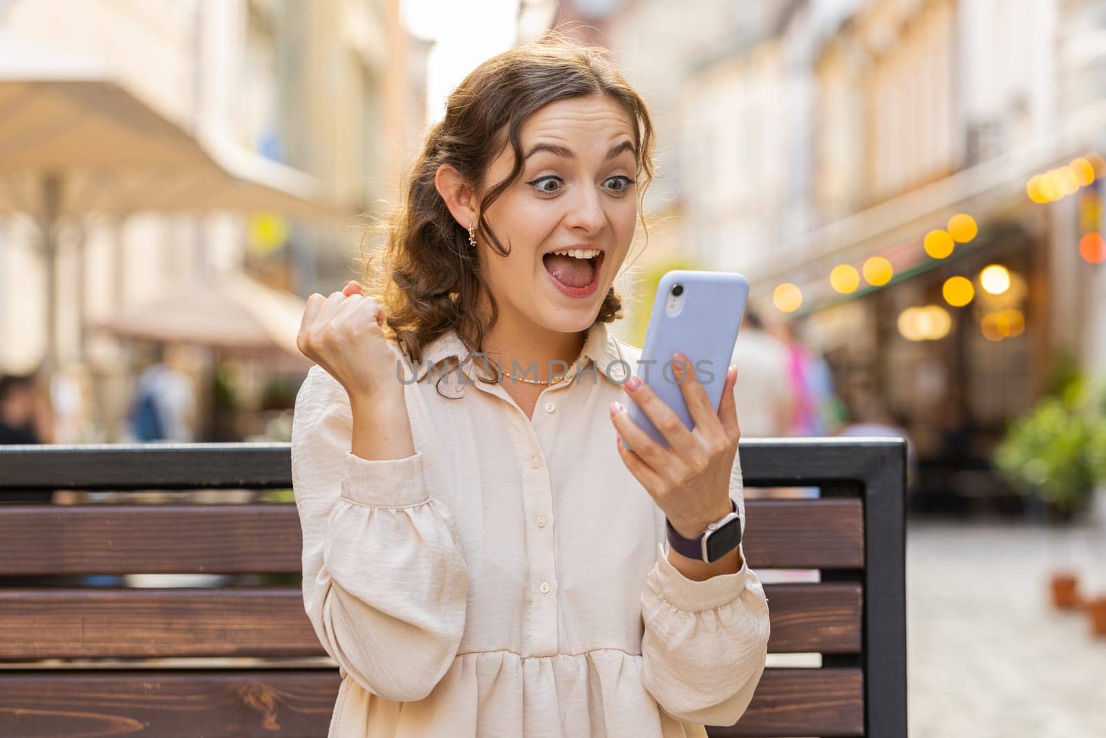 Amazed young pretty woman use mobile smartphone celebrating win good message news, lottery jackpot victory, giveaway online. Happy lovely girl sits on bench on city sunny street outdoors. Lifestyles
