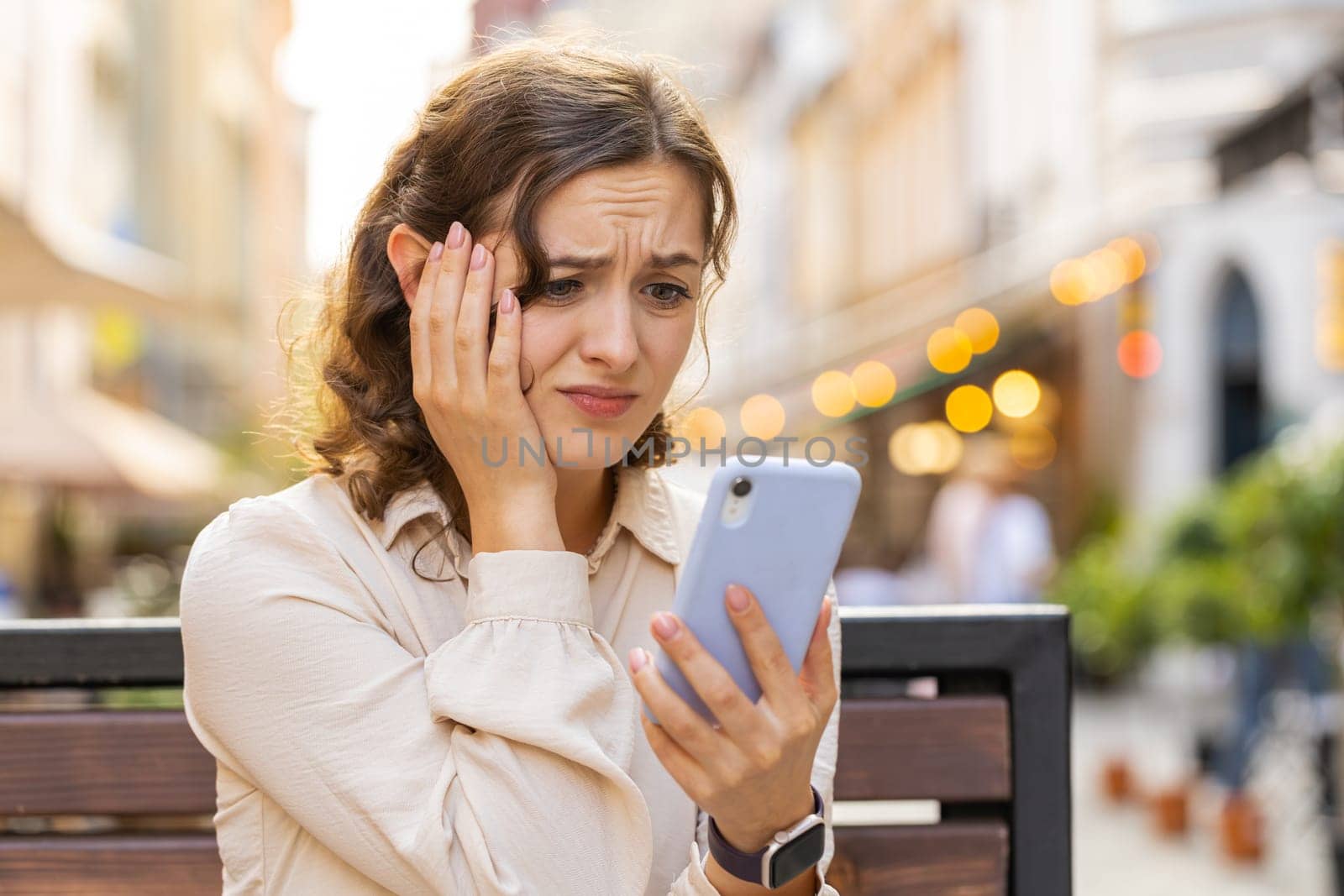 Young pretty woman use smartphone typing browsing, loses becoming surprised sudden lottery results, bad news, fortune loss, fail. Lovely girl sits on bench on city street outdoors. Town lifestyles