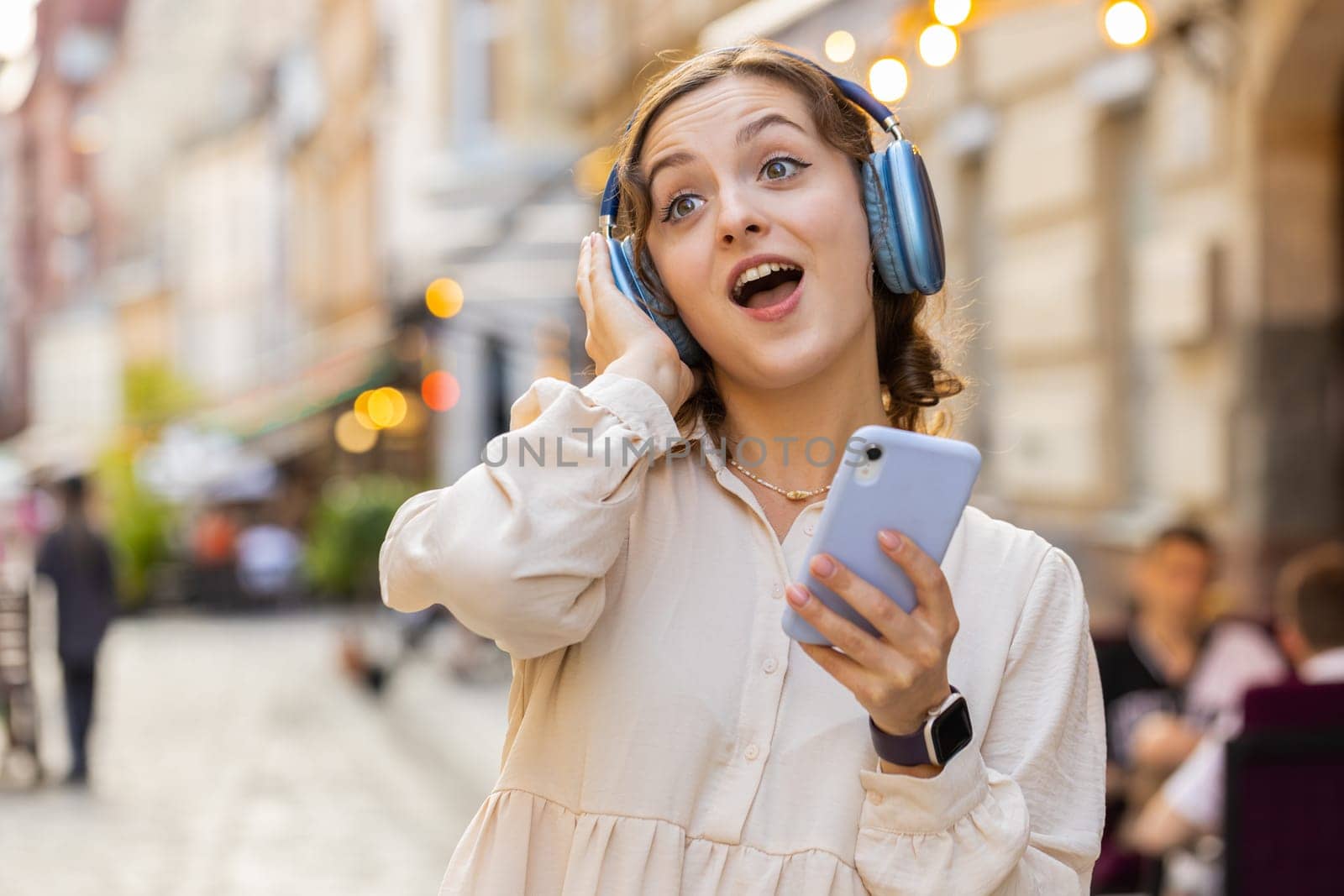Happy relaxed overjoyed pretty young woman in wireless headphones choosing, listening favorite energetic disco rock n roll music dancing outdoors. Girl walking in urban city sunshine street background